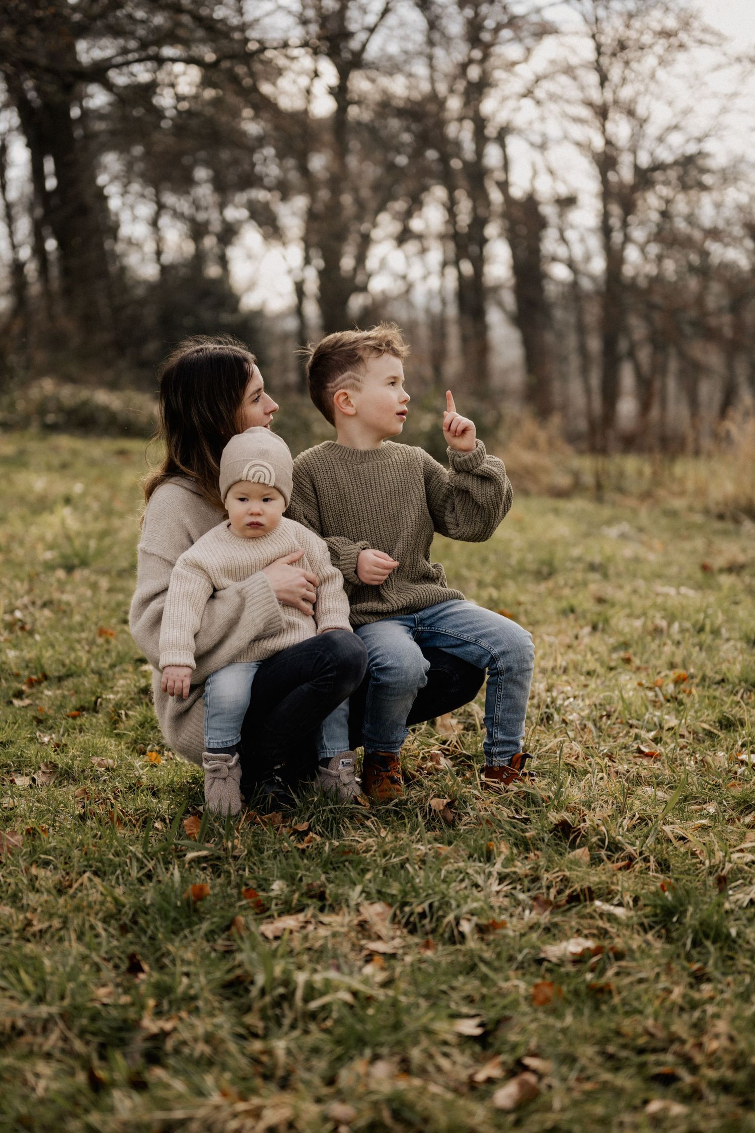 A cozy family moment on a grassy hillside surrounded by bare trees in soft muted tones.