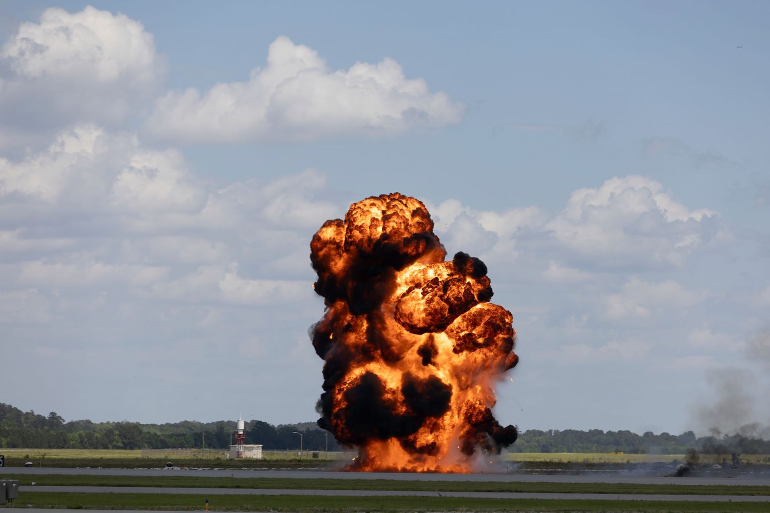 Large explosion with orange fireball and black smoke on airfield during military demonstration.