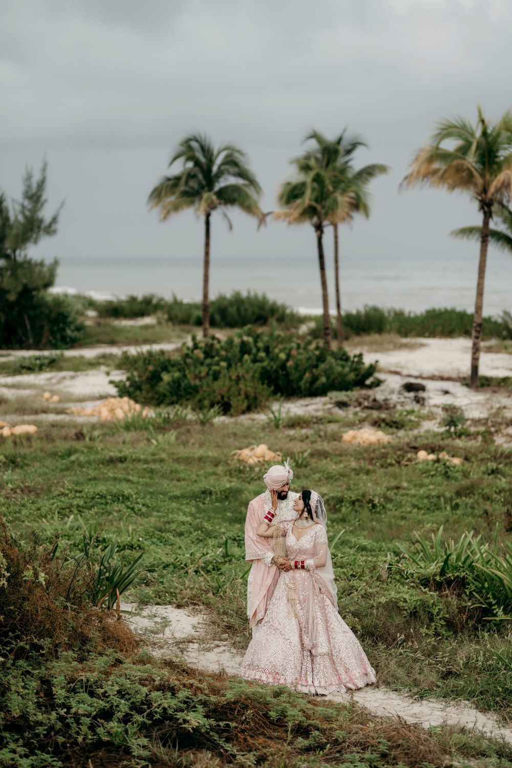 A couple in romantic pink attire embrace on a tropical beach path with palm trees and cloudy skies in the background.