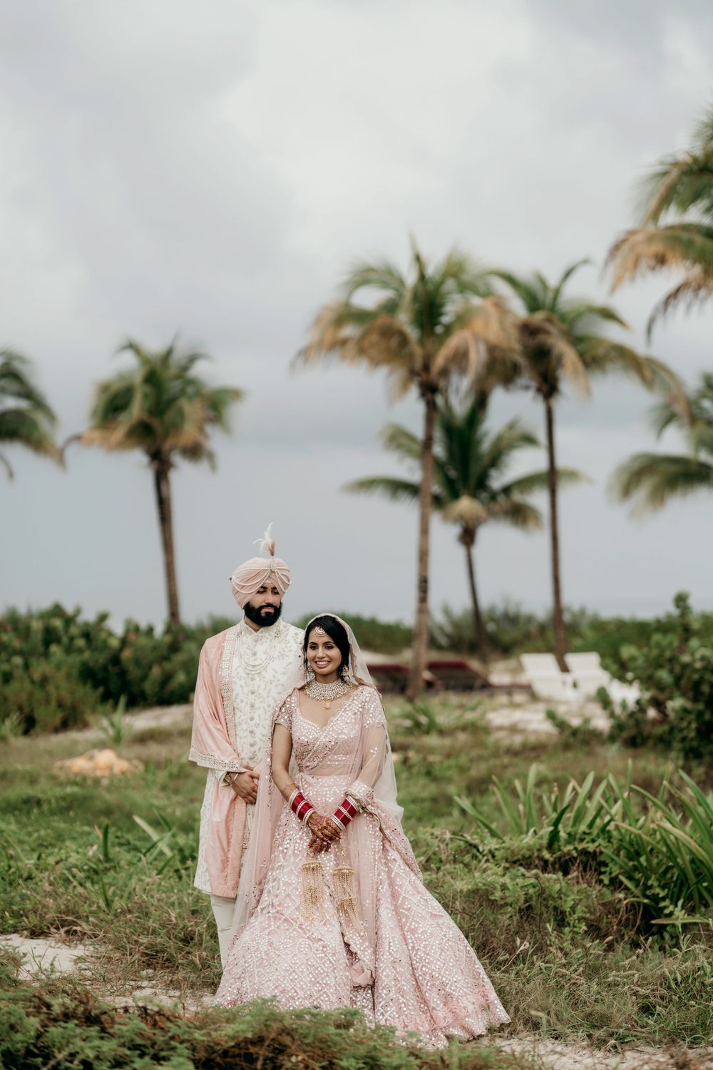 Traditional wedding couple in pink attire poses together on a tropical beach with palm trees and cloudy sky.