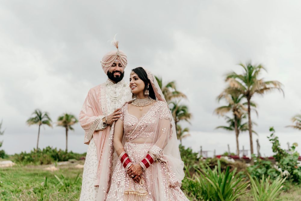A couple in traditional pink ornate Indian wedding attire pose together outdoors with palm trees in the background.
