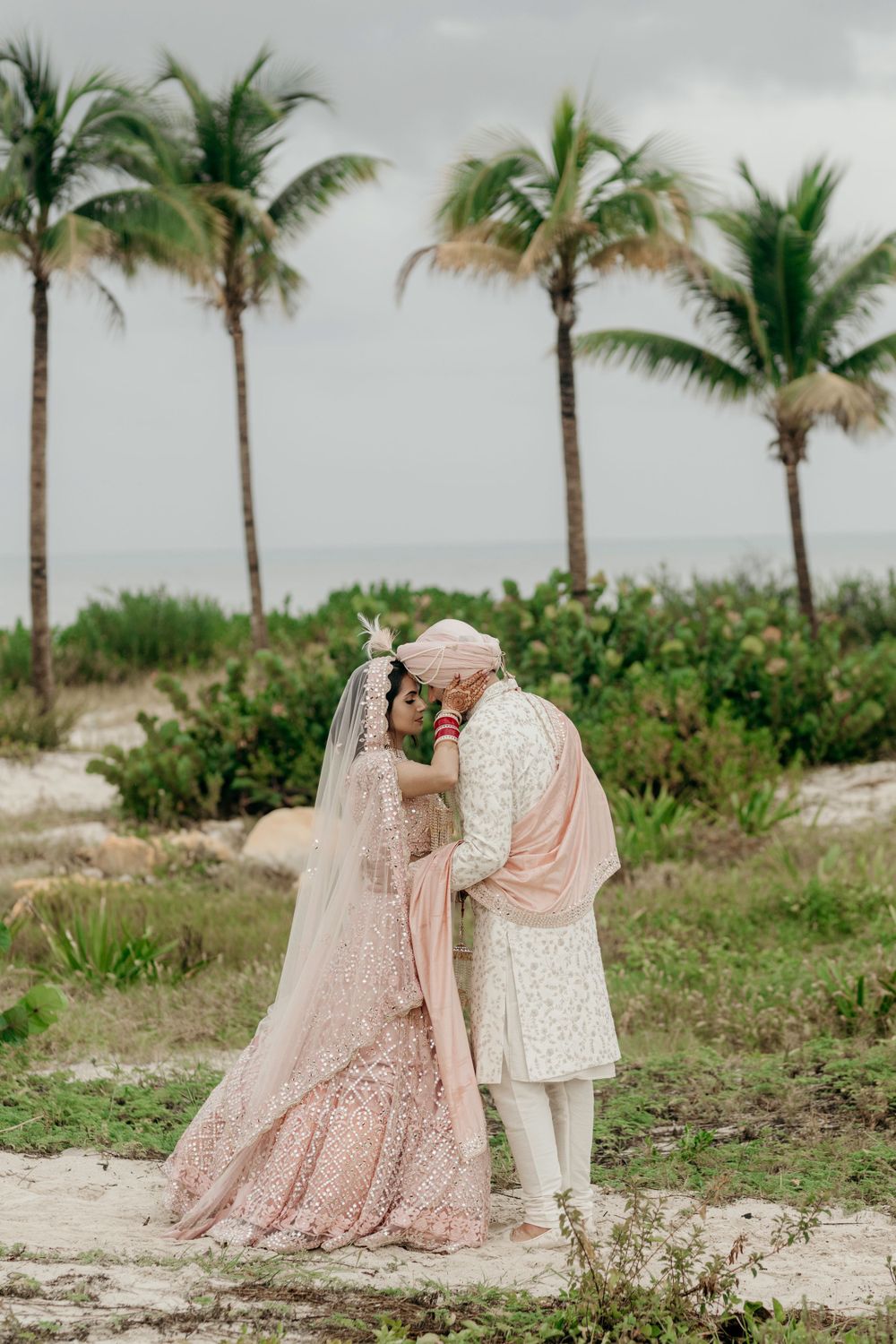 A couple in traditional South Asian wedding attire embrace on a tropical beach with palm trees in the background.
