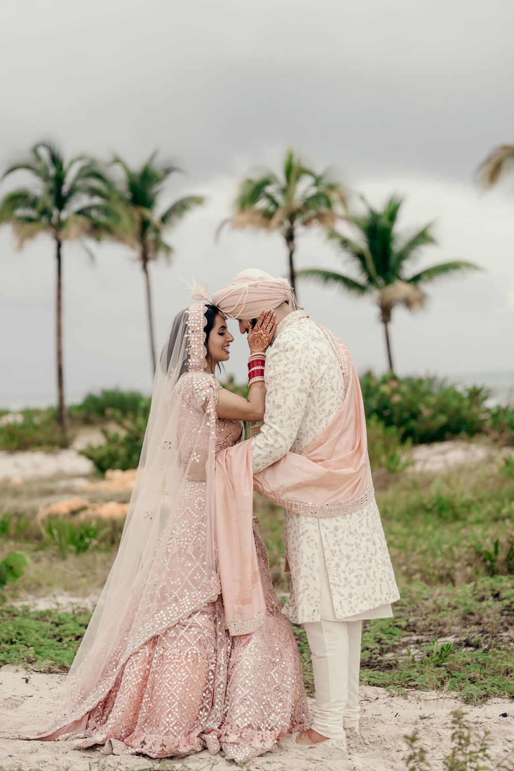 A romantic couple in traditional South Asian wedding attire embrace on a beach with palm trees in the background.