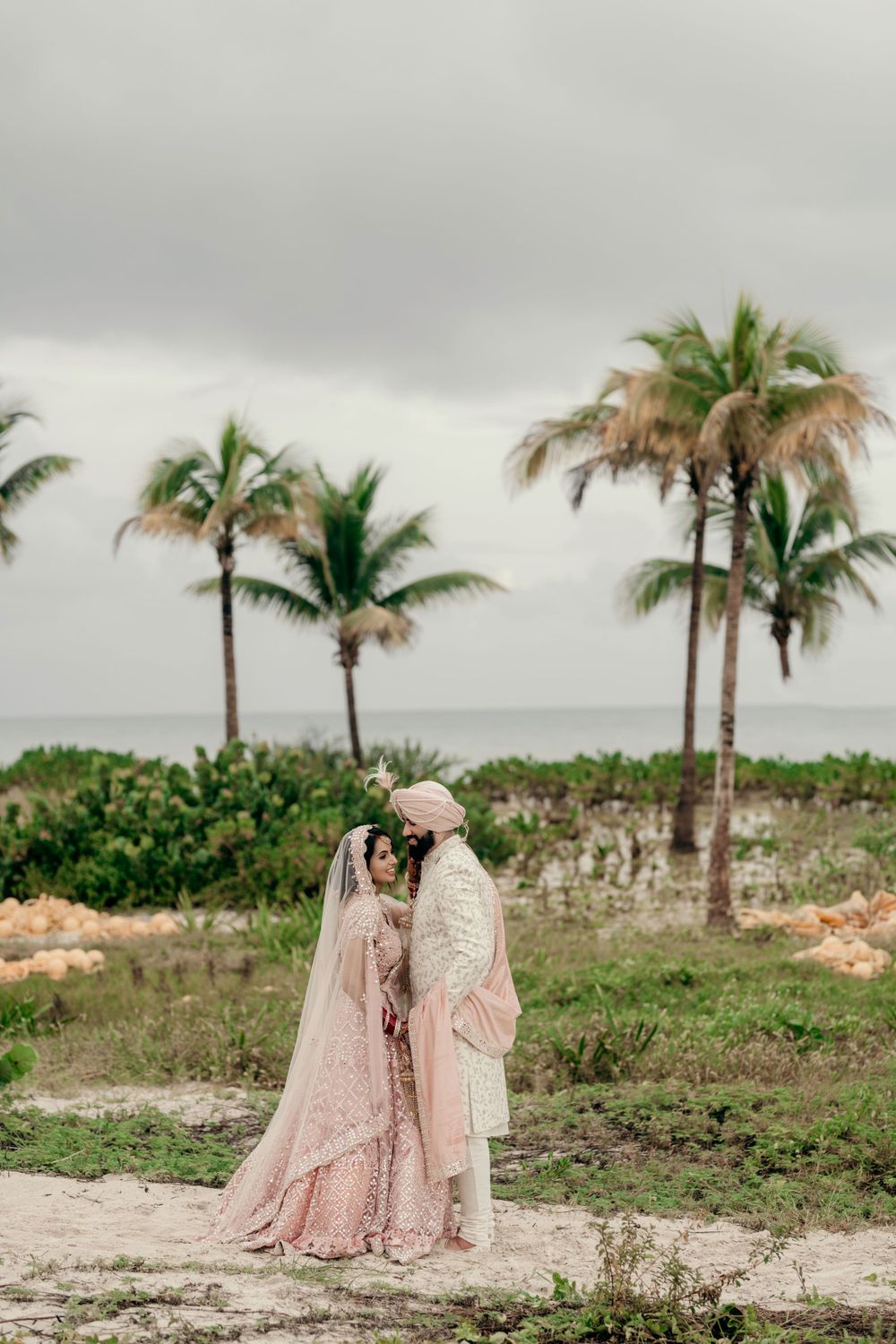 A couple in traditional South Asian wedding attire stands on a beach with palm trees and cloudy skies in the background.