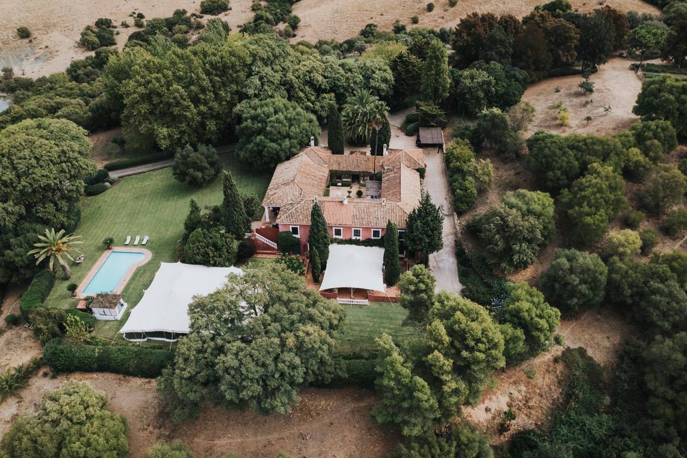 Aerial view of a rustic estate with swimming pool surrounded by lush gardens and trees in a countryside setting.