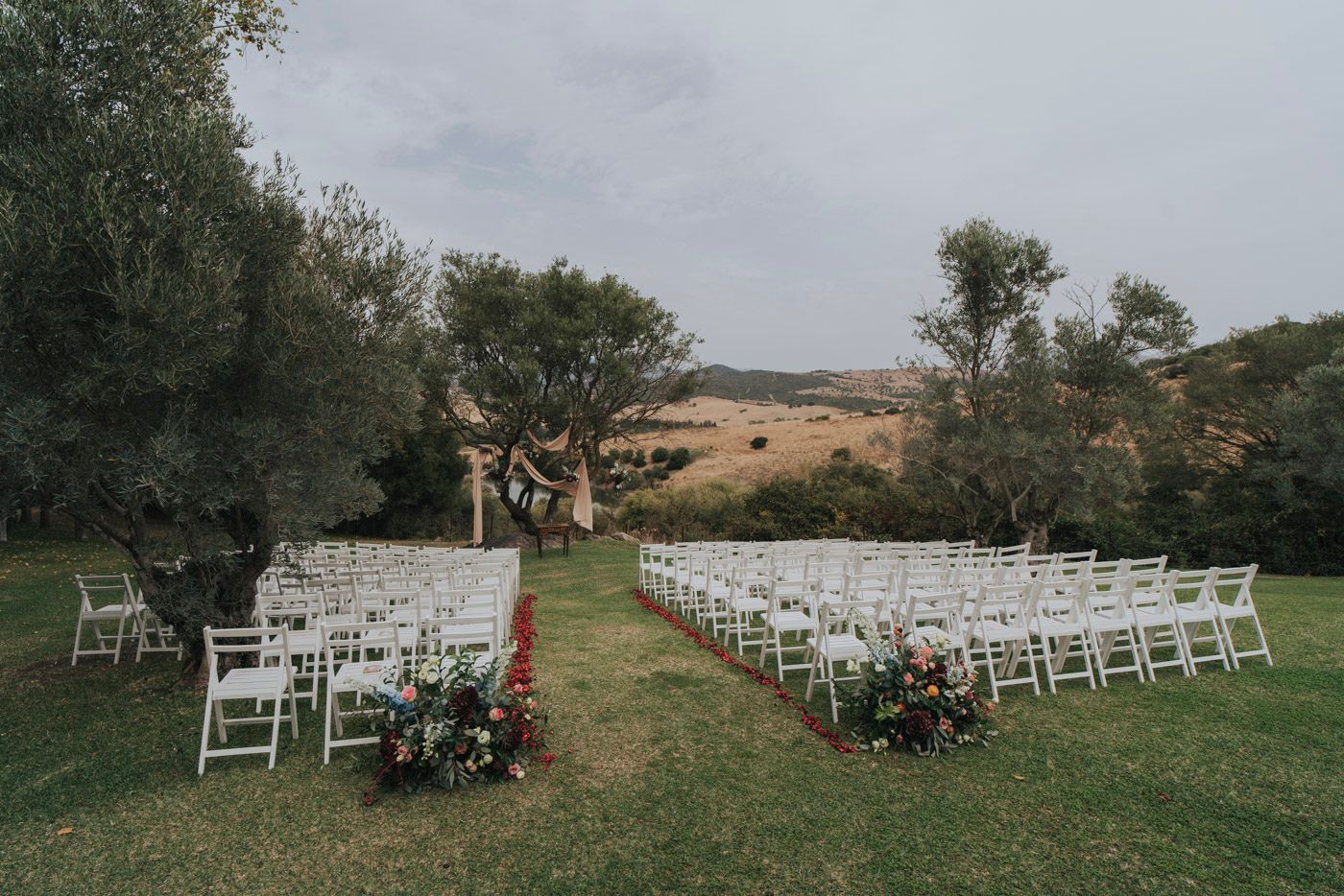 White wedding chairs arranged in rows on a grassy hillside with olive trees and flower arrangements in an outdoor venue setup.