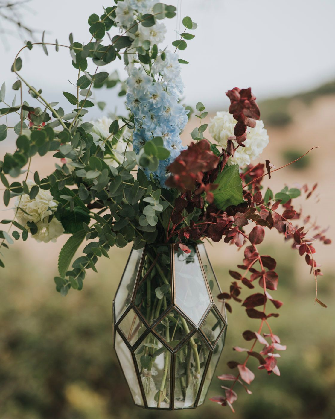 Delicate floral arrangement with eucalyptus and burgundy blooms in a geometric glass vase against a soft blurred background.