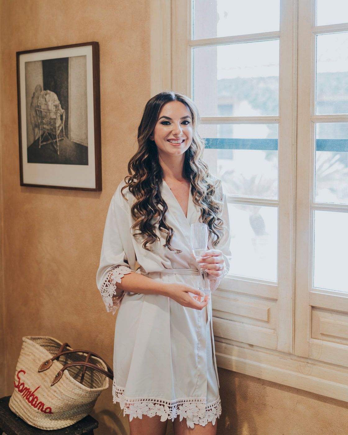 Someone wearing a white lace-trimmed robe stands by a window in a room with warm beige walls and decorative artwork.