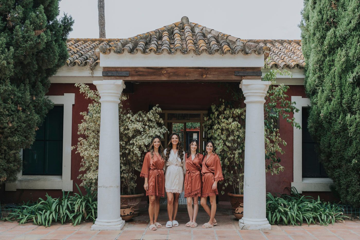 Bridal party poses under Spanish-style archway with white columns and terracotta roof at elegant garden venue.