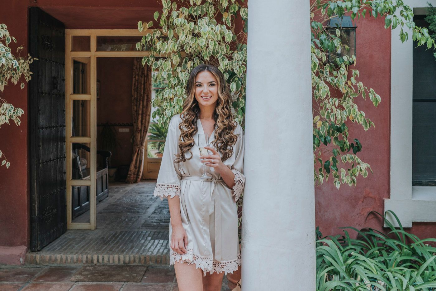 Woman in white lace robe standing by column with climbing vines against pink wall.