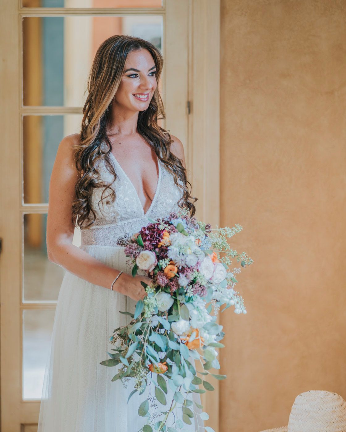 Wedding photo showing elegant white bridal gown and cascading pastel floral bouquet against adobe wall and french doors.