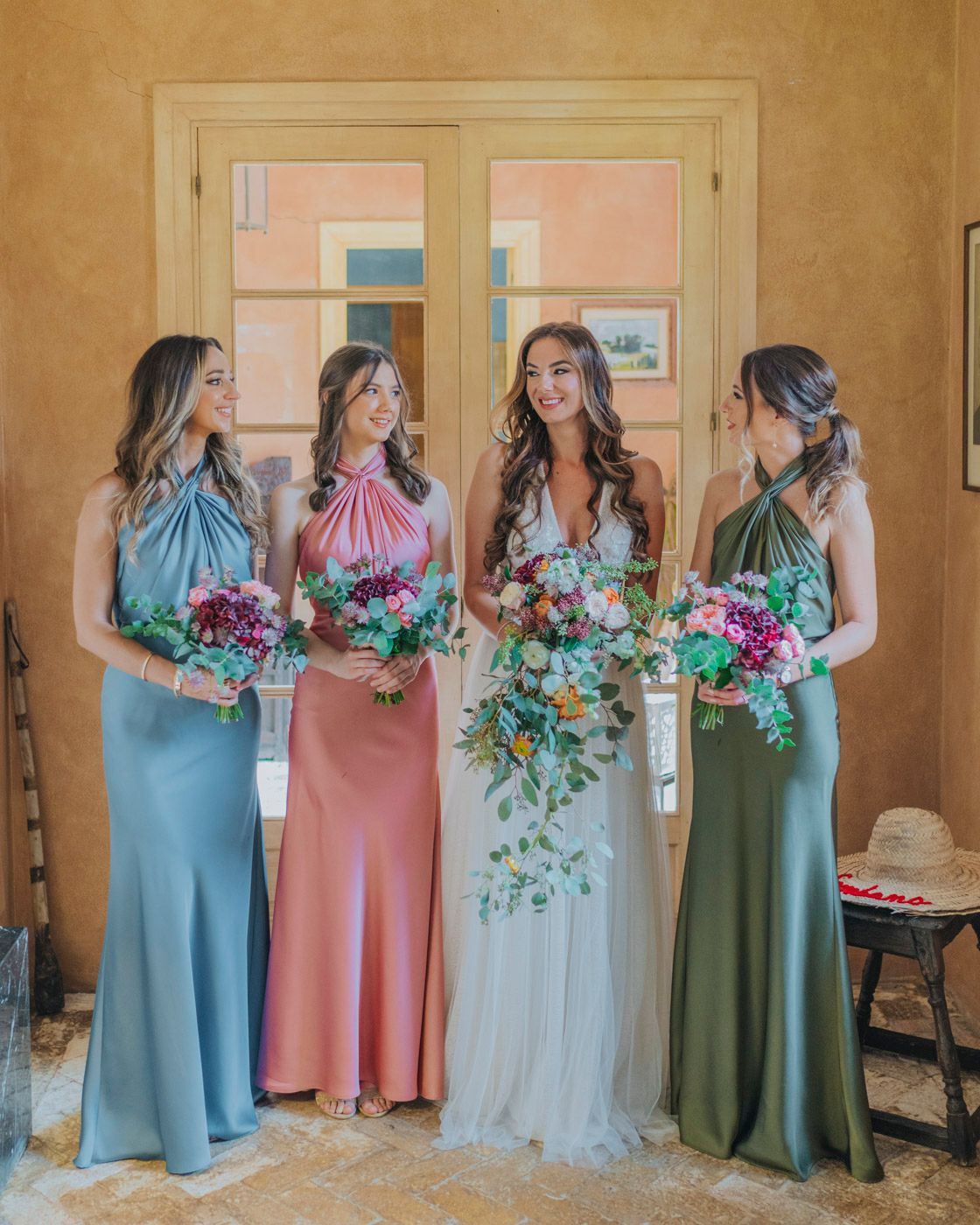 Bridesmaids in jewel-toned dresses pose with bride wearing white gown, all holding colorful floral bouquets.