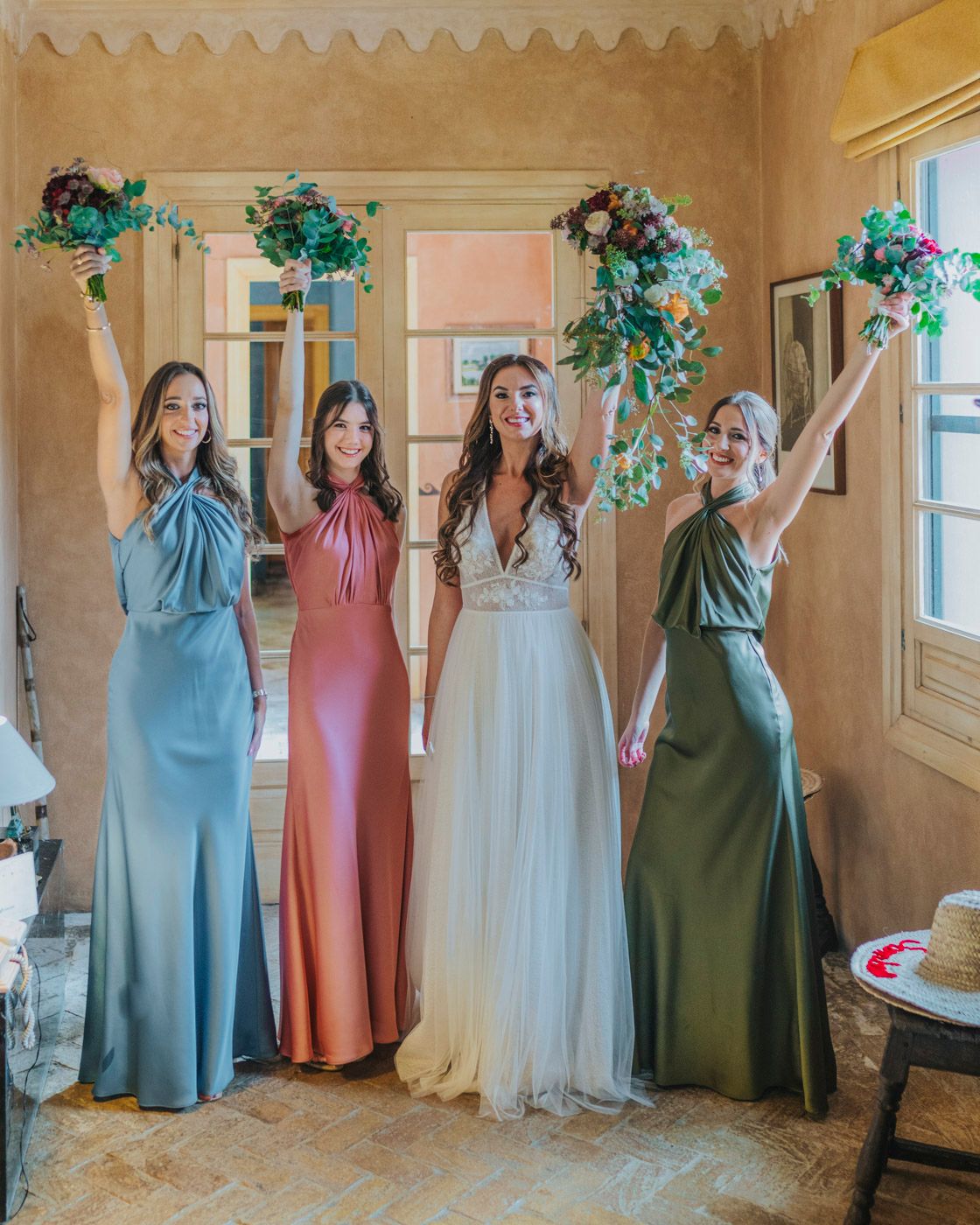 Wedding party strikes fun pose against adobe wall while holding vibrant bouquets in coordinating pastel dresses.