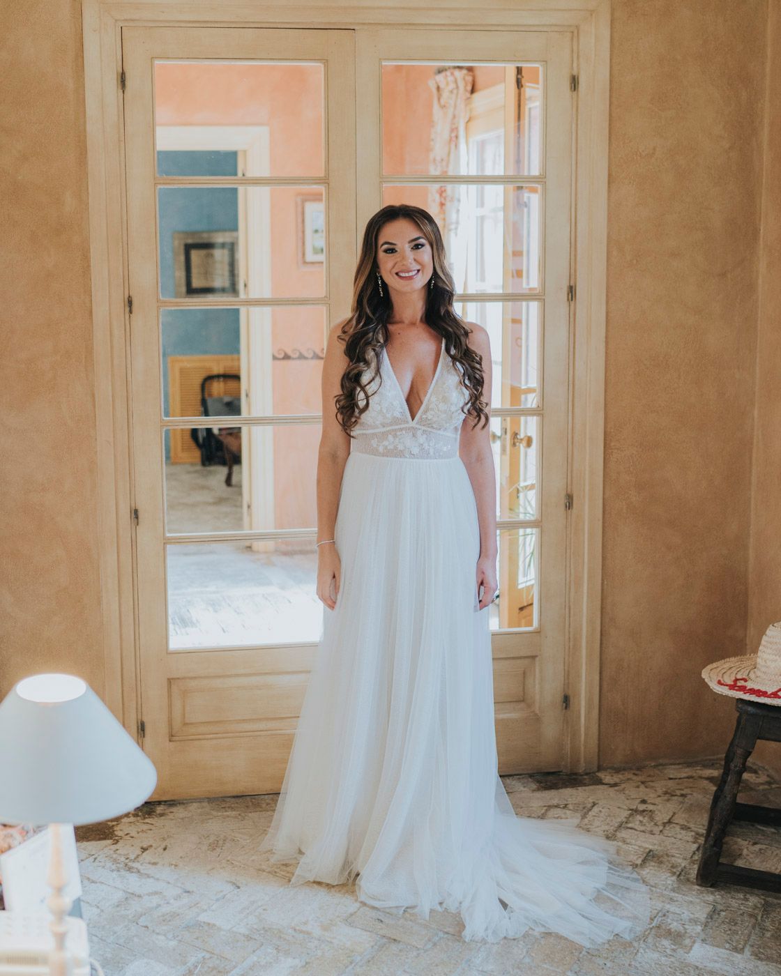 A flowing white wedding gown with lace details and deep v-neckline is showcased in an elegant interior setting.