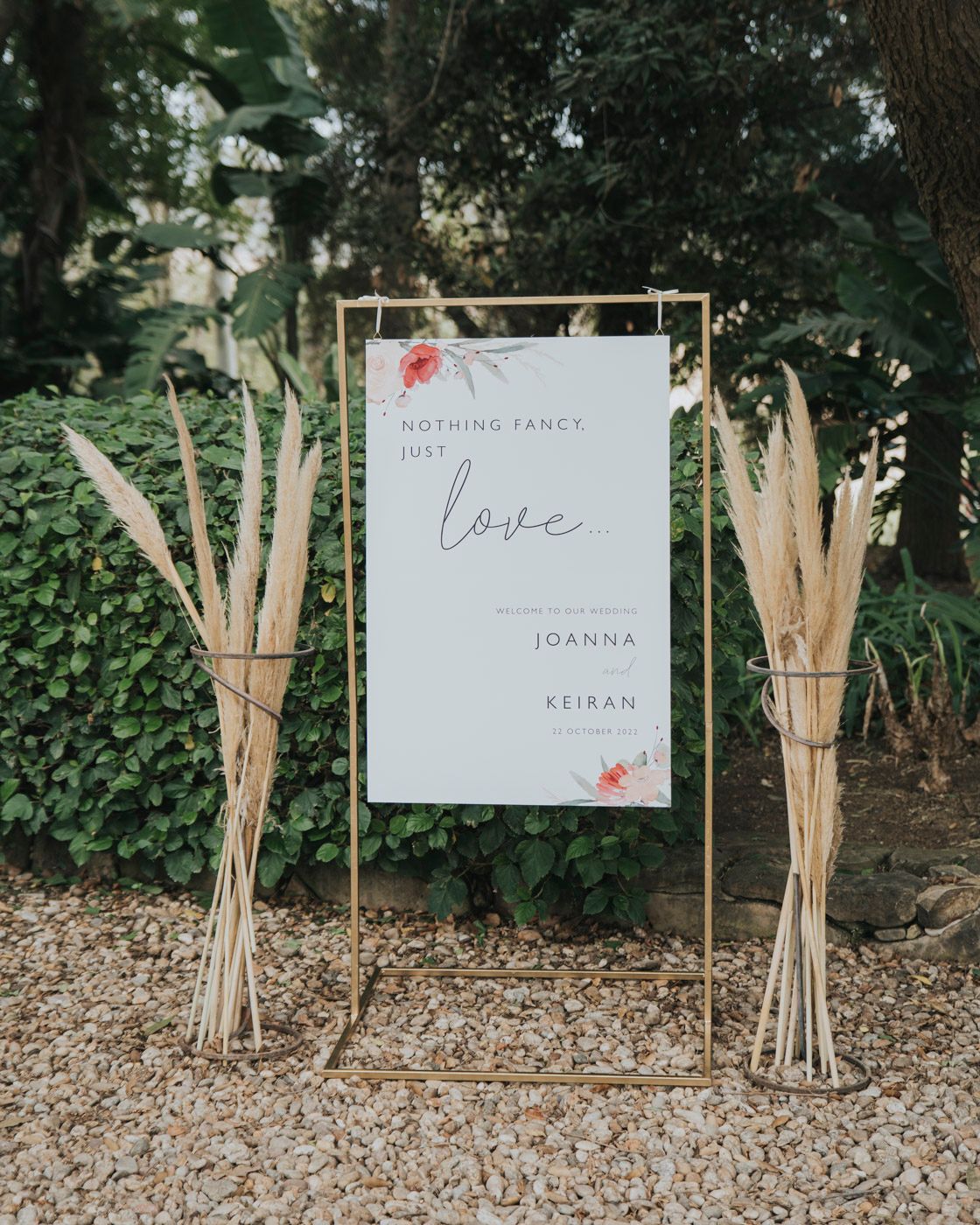 A wedding welcome sign with floral accents displayed between two dried pampas grass arrangements on a garden path.