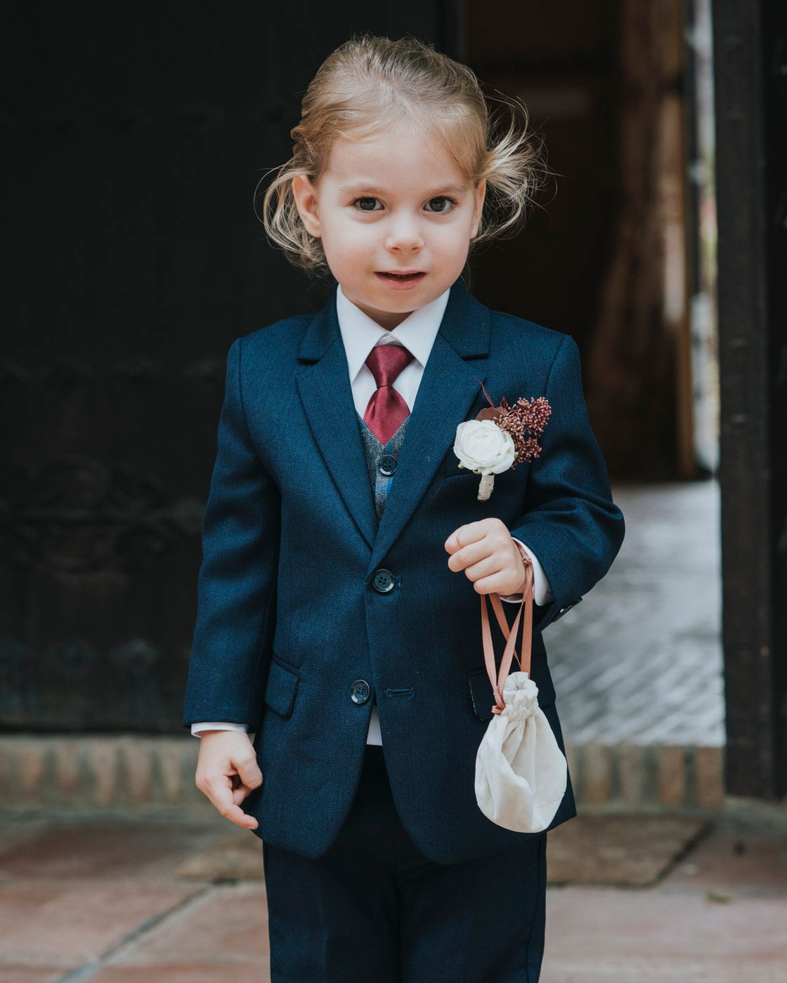 A young child in a navy blue suit with red tie and boutonniere carries a small white drawstring bag at a formal event.