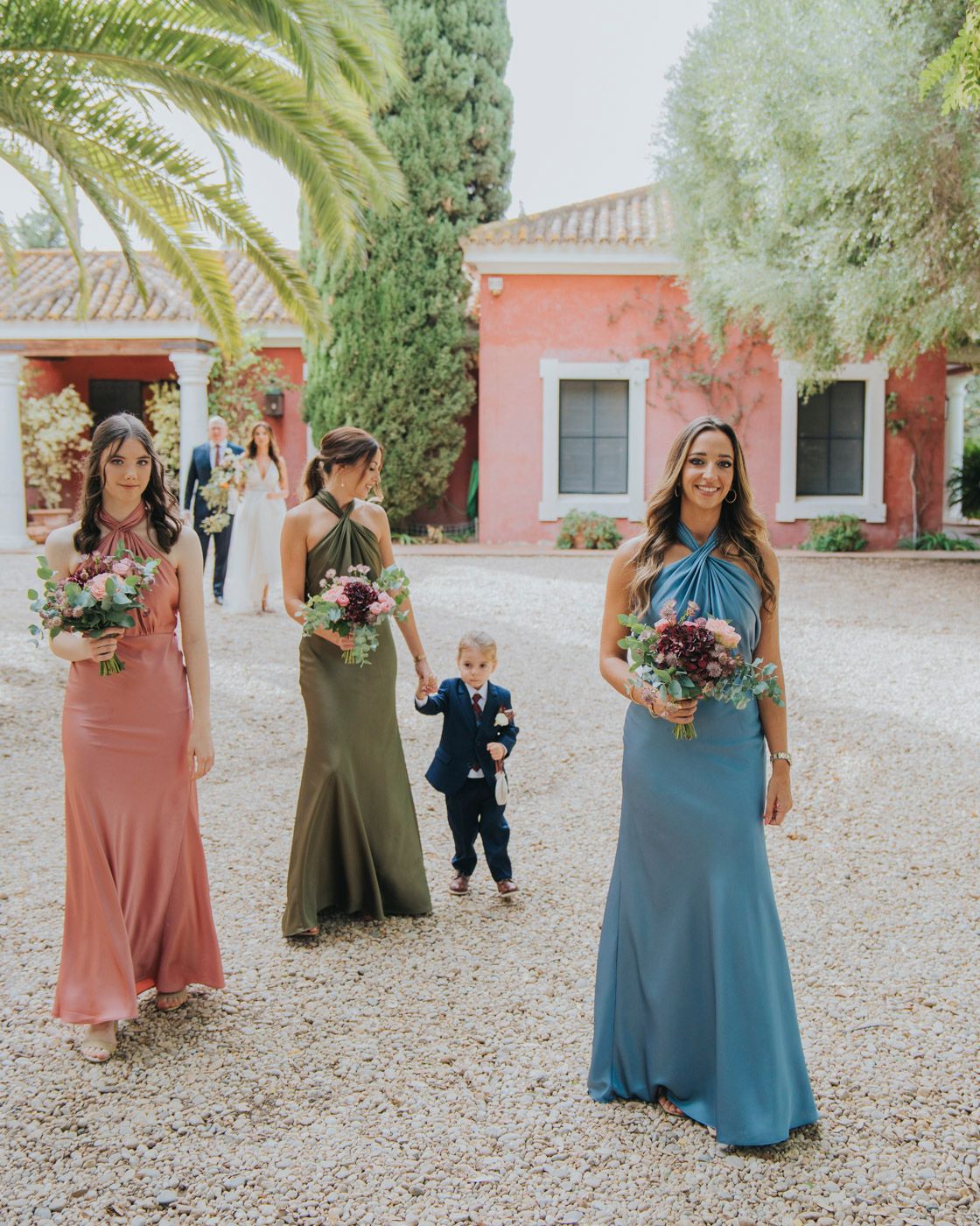 Wedding party members in flowing dresses stand on a gravel path by a Mediterranean-style villa with palm trees and cypress.
