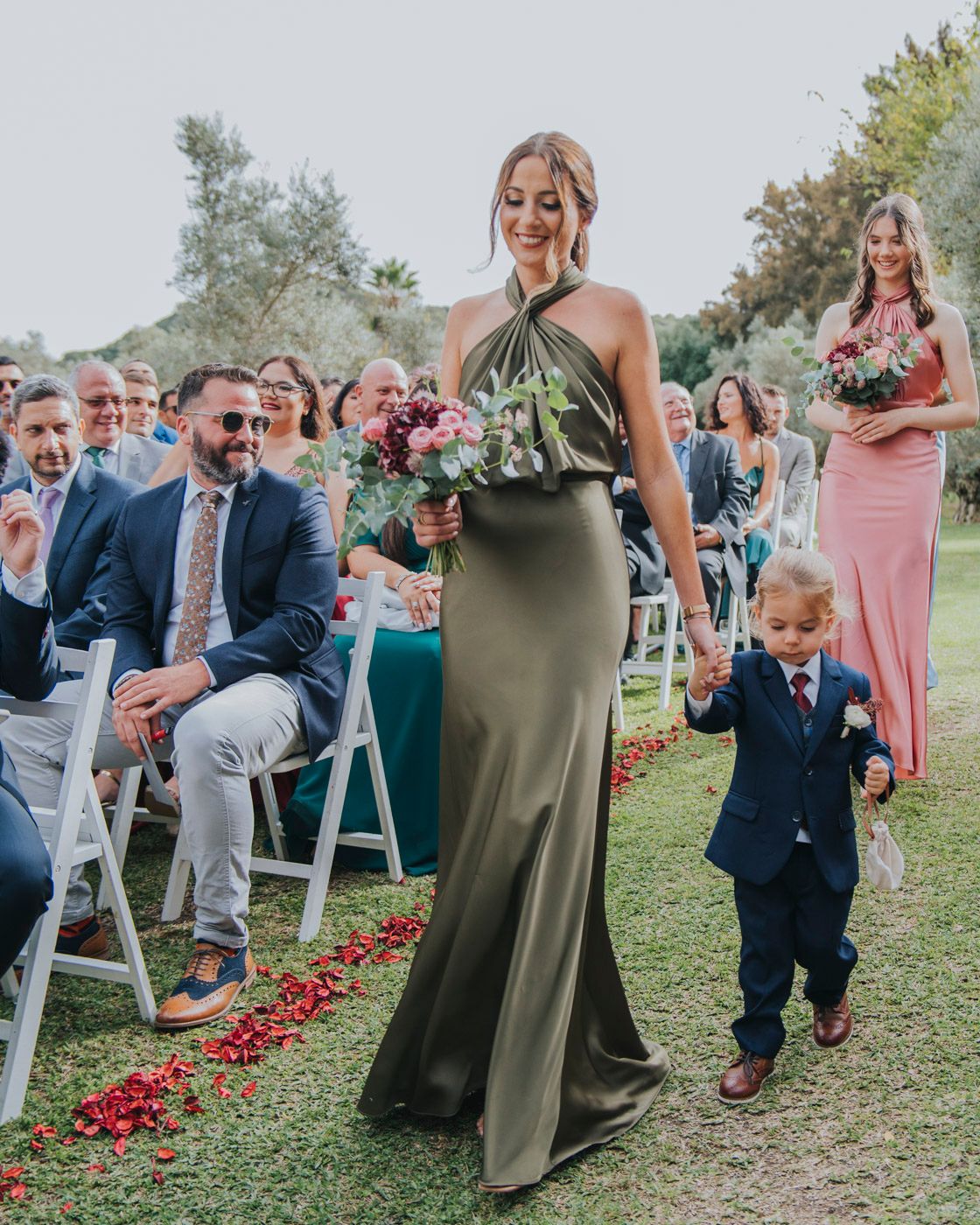 Wedding attendees and bridal party walk down a rose petal-lined aisle during an outdoor garden ceremony.