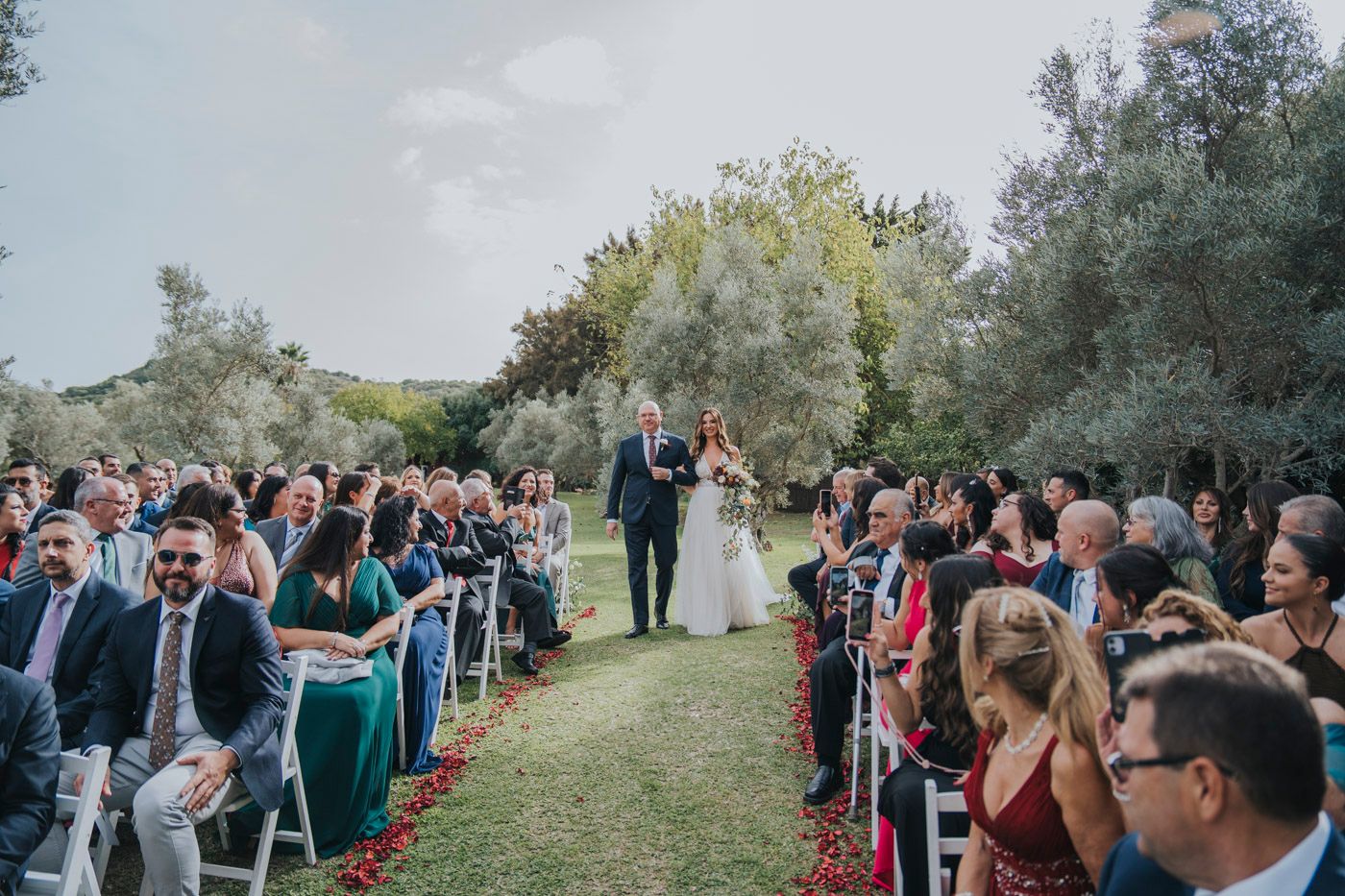 An outdoor wedding ceremony on a grassy path surrounded by guests seated in white chairs under green trees.