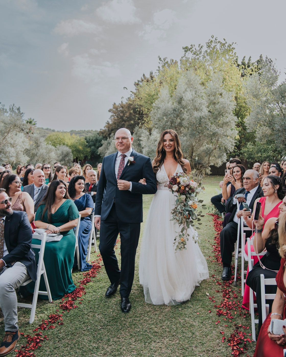 An outdoor wedding ceremony with rose petals lining the aisle as a couple proceeds between seated guests on the lawn.