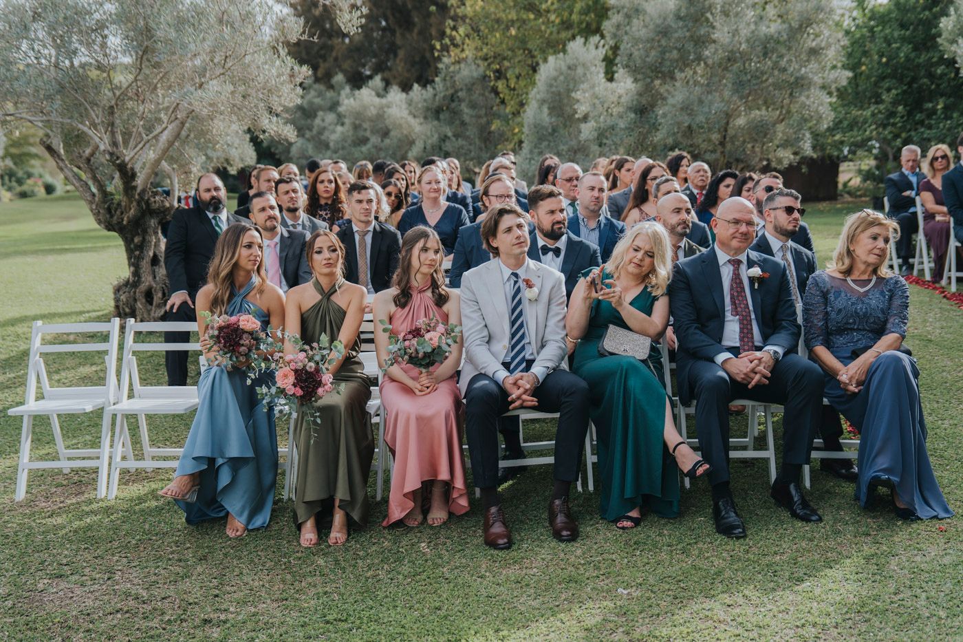 Wedding guests seated in white chairs on a green lawn during an outdoor ceremony on a sunny day.