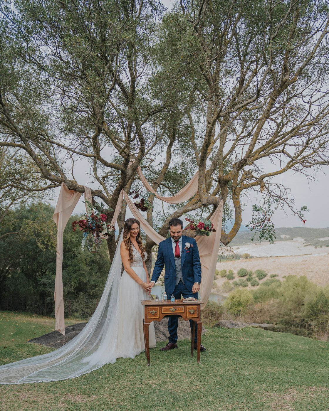 A couple celebrates their wedding ceremony under draped fabric and trees at an outdoor coastal venue on a grassy hillside.