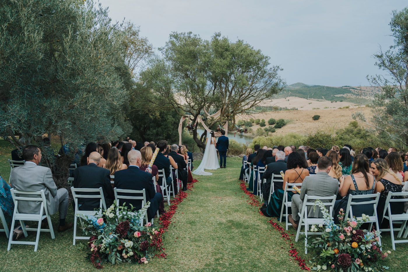 An outdoor wedding ceremony setup with white folding chairs and floral arrangements along a grassy aisle in a scenic countryside.