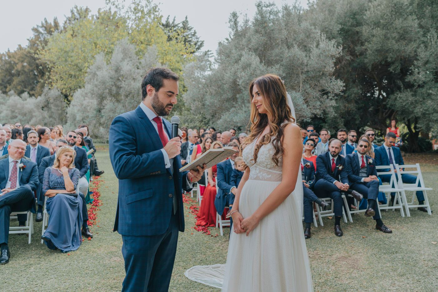 A couple exchanging vows at an outdoor wedding ceremony with guests seated on white chairs in a garden setting.