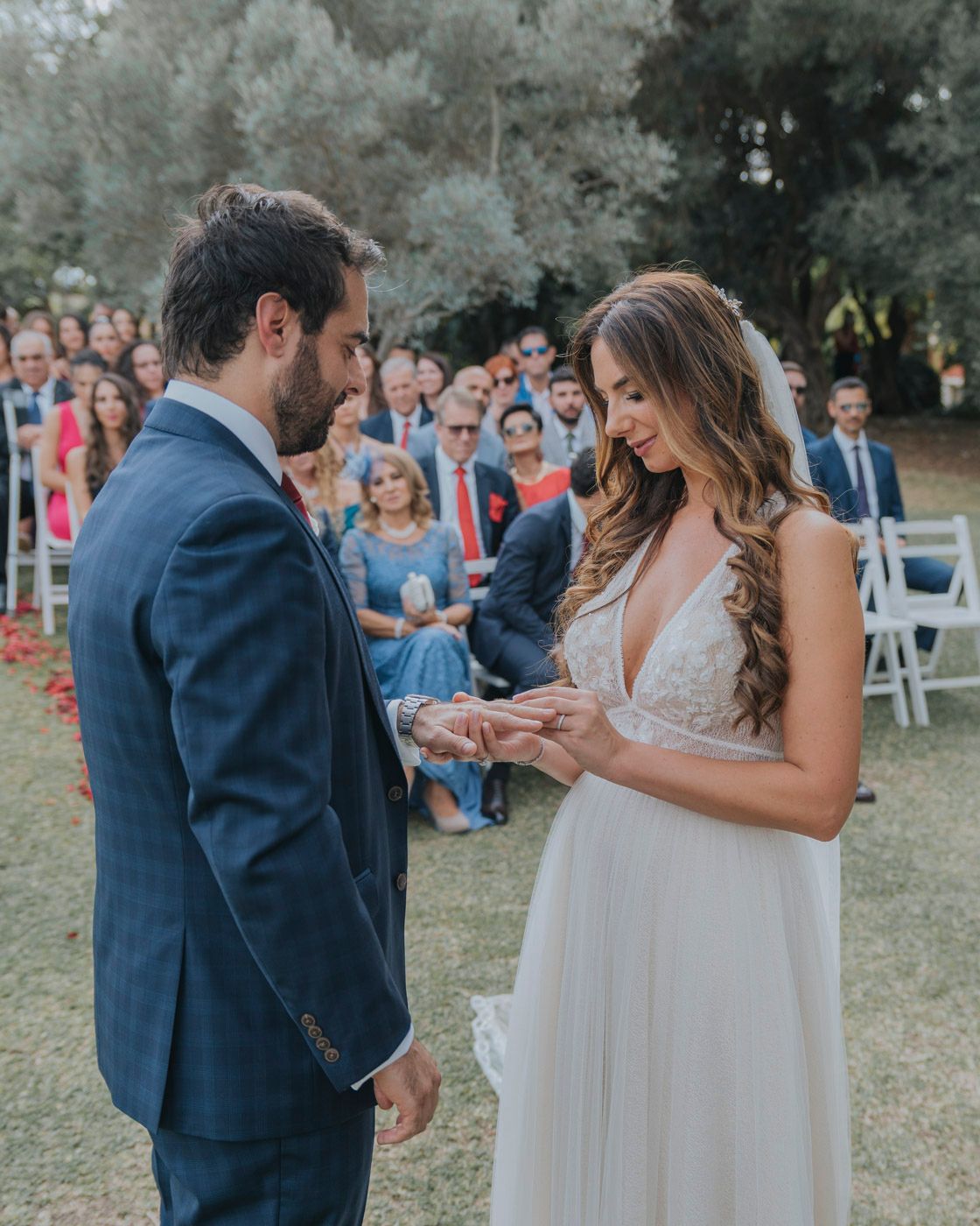 Couple exchanging rings during an outdoor wedding ceremony while guests watch from white chairs in a garden setting.