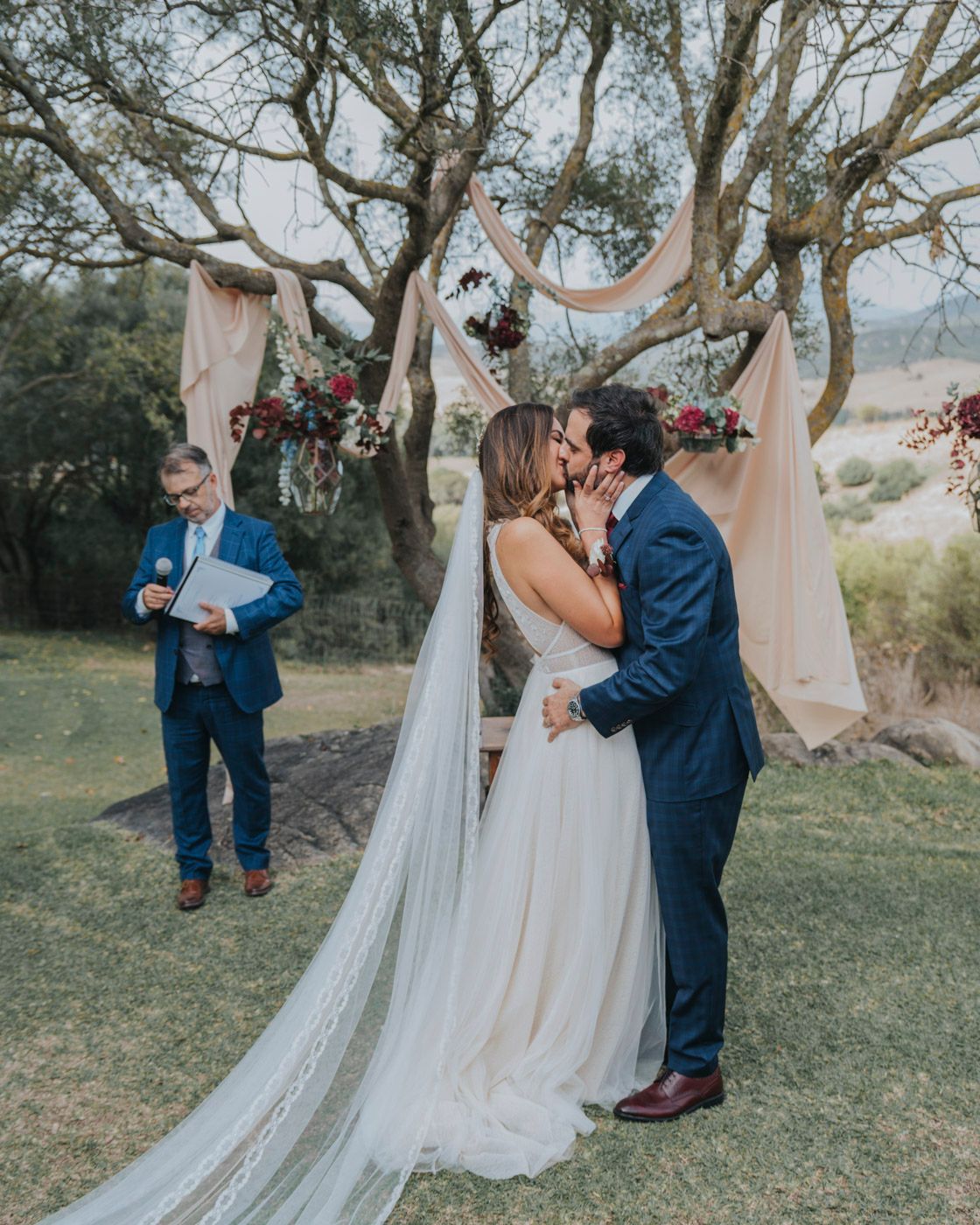 A wedding couple sharing a romantic moment under a blush-draped arch with floral arrangements in a garden.