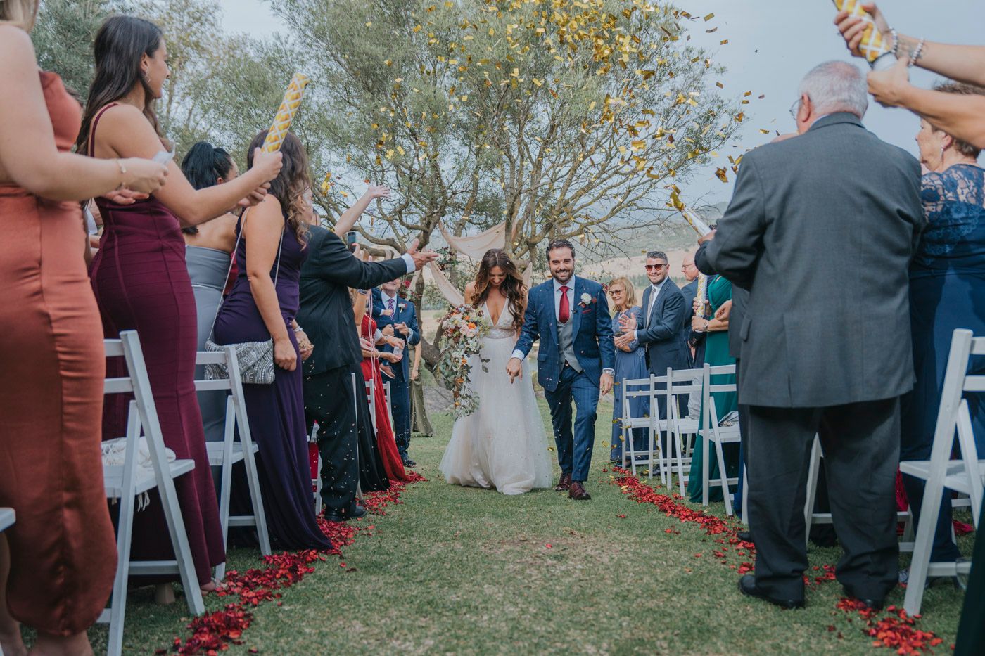Wedding guests line an outdoor aisle with wands and ribbons as a couple walks between them during a ceremony.