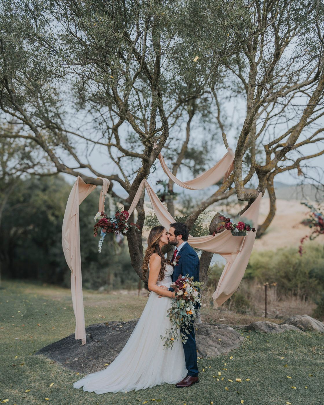 A romantic wedding ceremony setup with flowing fabric draping from olive trees decorated with burgundy floral arrangements.