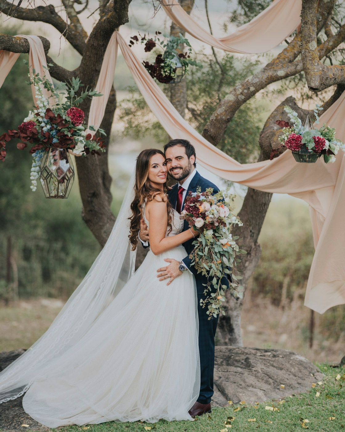 Romantic outdoor wedding scene with flowing blush drapery, floral arrangements and lanterns decorating tree branches in nature.