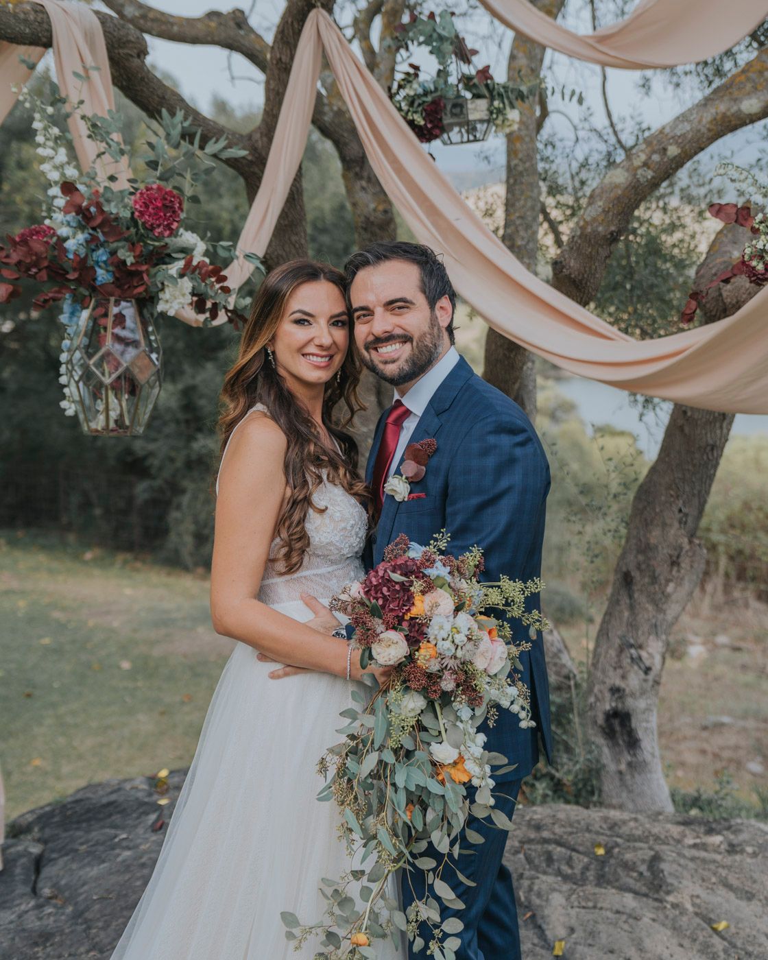 Romantic outdoor wedding celebration with floral arrangements and blush drapery decorating natural tree branches in garden setting.