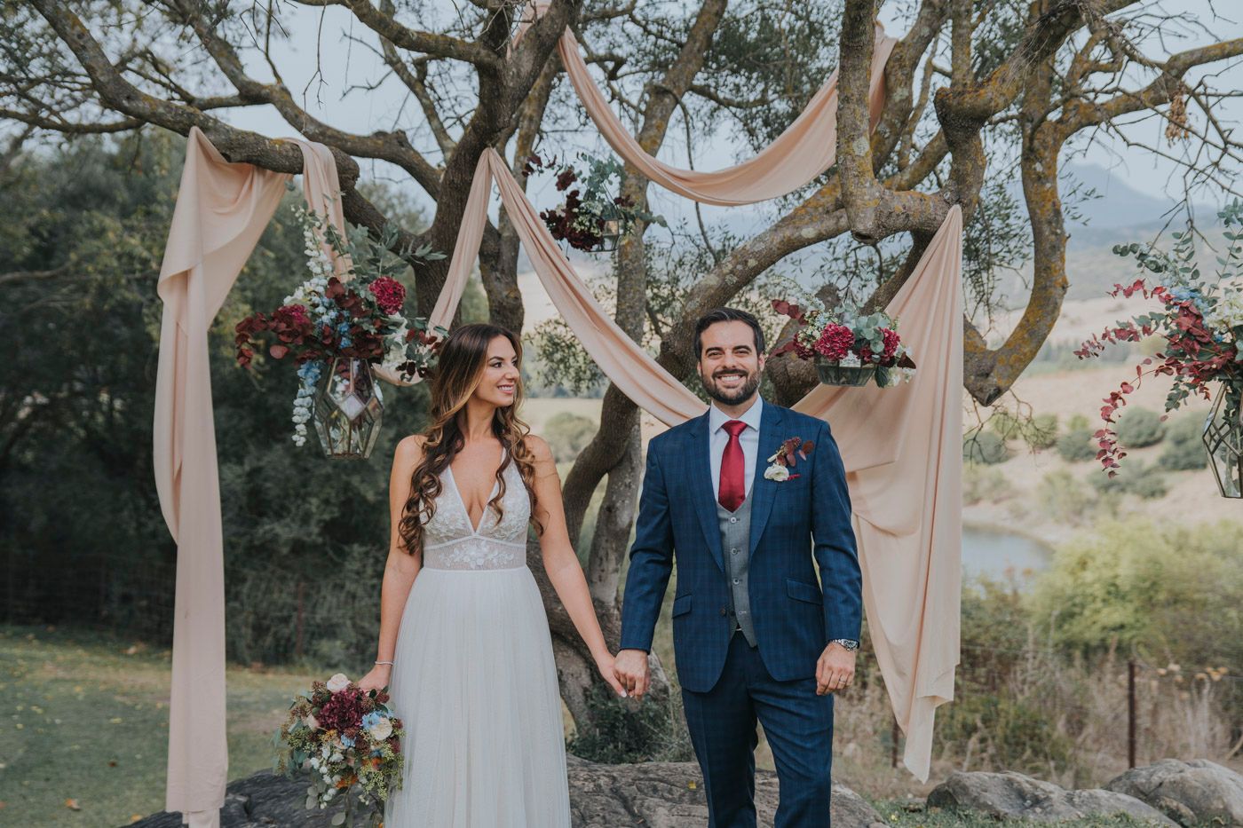 A rustic outdoor wedding ceremony setup with draped blush fabric and floral decorations under old oak trees.