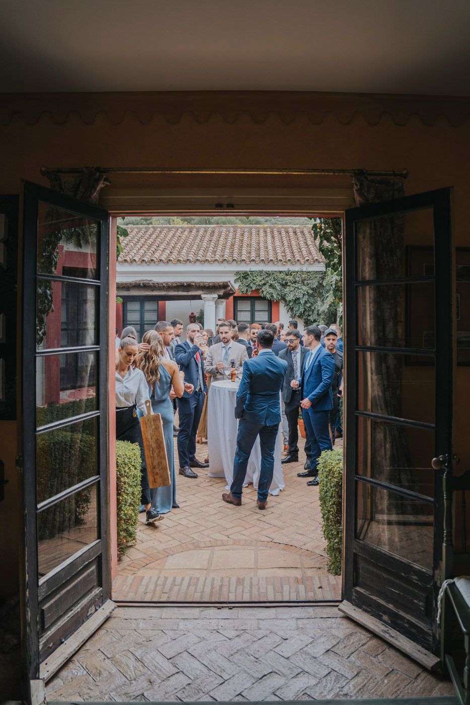 View through open French doors of guests gathered in a courtyard with traditional Chinese architecture and manicured hedges.