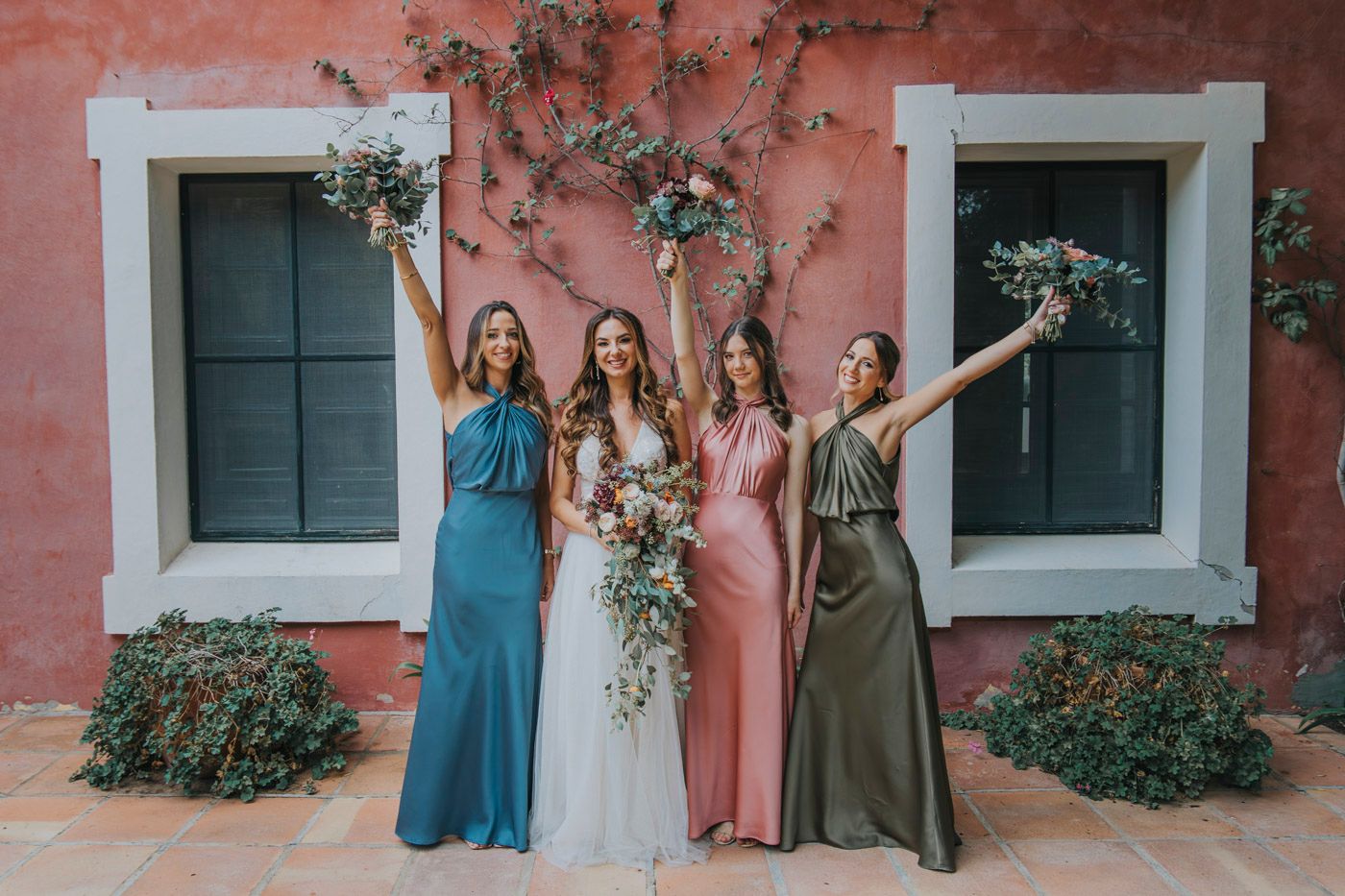 Wedding party group stands in front of coral colored wall with green vines and two windows.