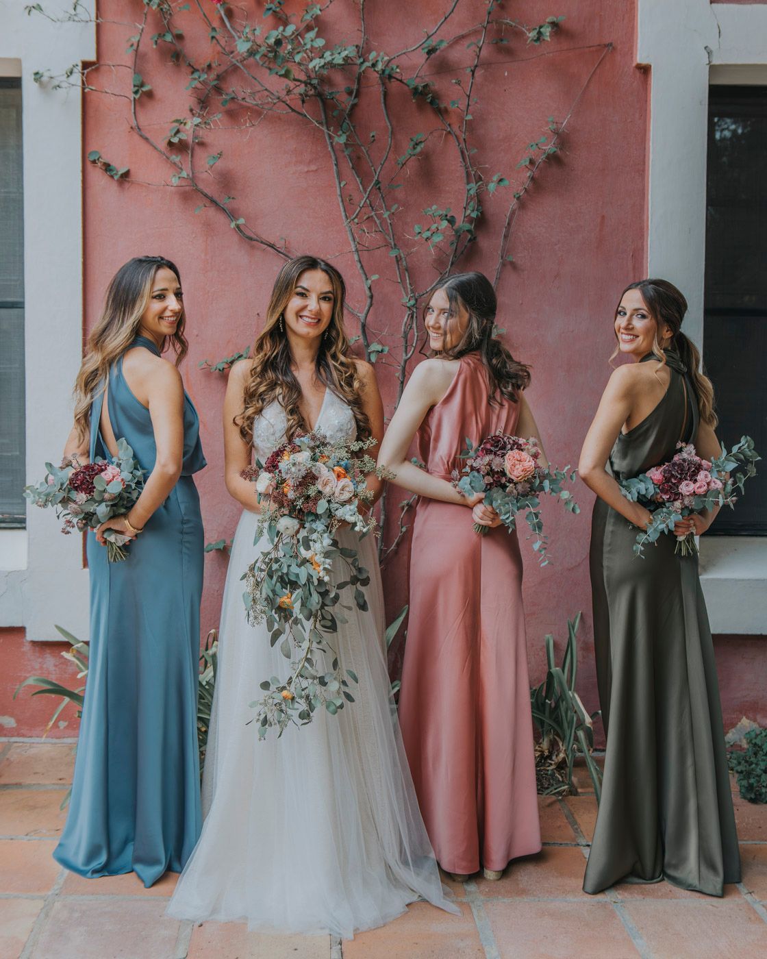 Wedding party in coordinating dresses of blue, white, pink and green pose against coral wall with climbing vines.