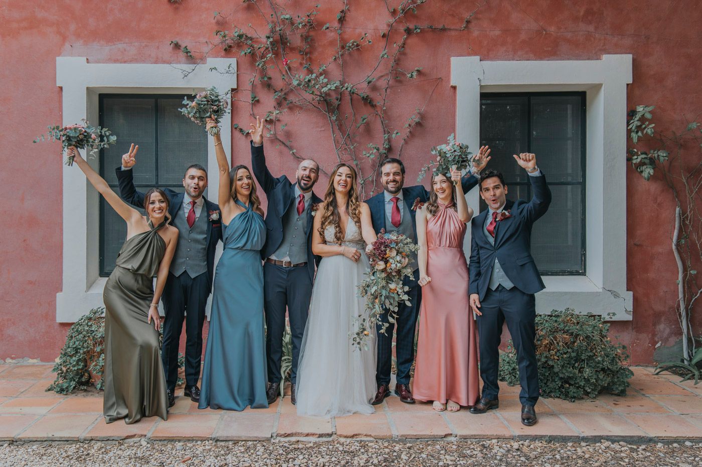 Celebratory group poses with raised arms against coral wall and windows at wedding celebration.