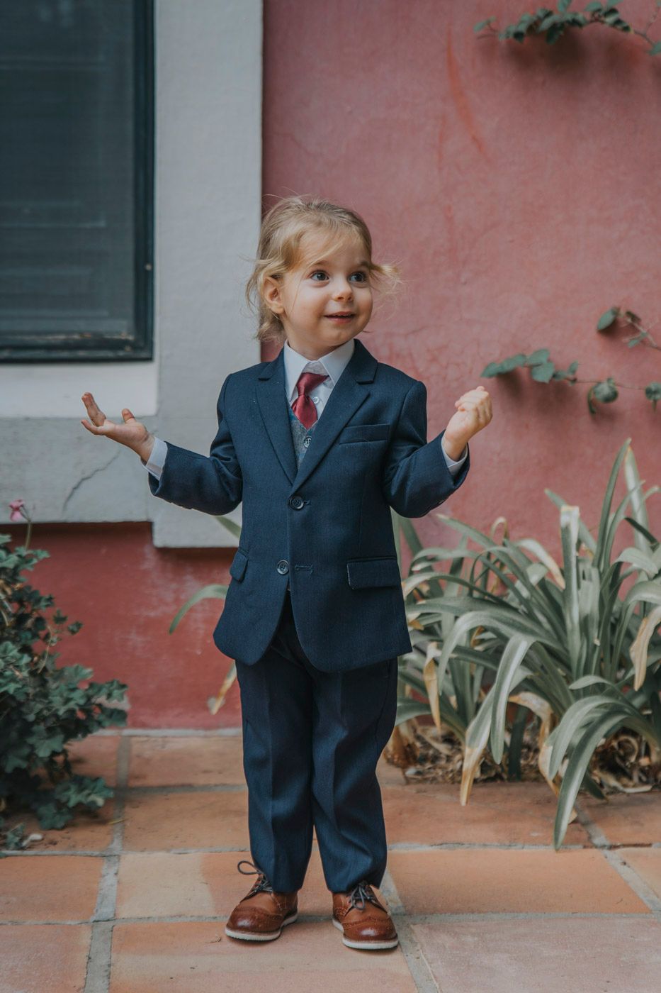 A young child in a navy suit and red tie gestures with their hands while standing against a coral-colored wall.