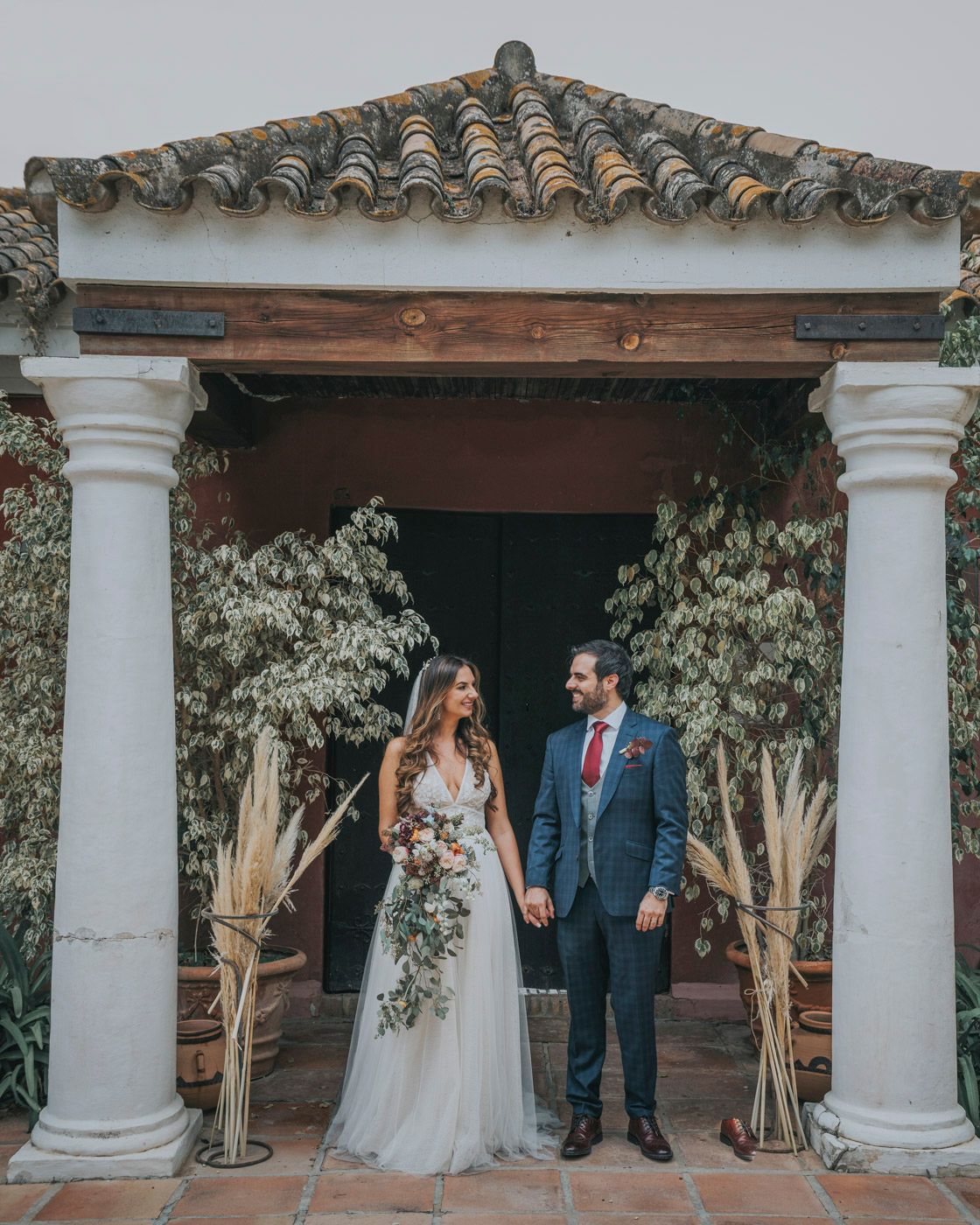 A wedding couple stands under a Spanish-style archway with white columns and terracotta roof tiles in a garden setting.