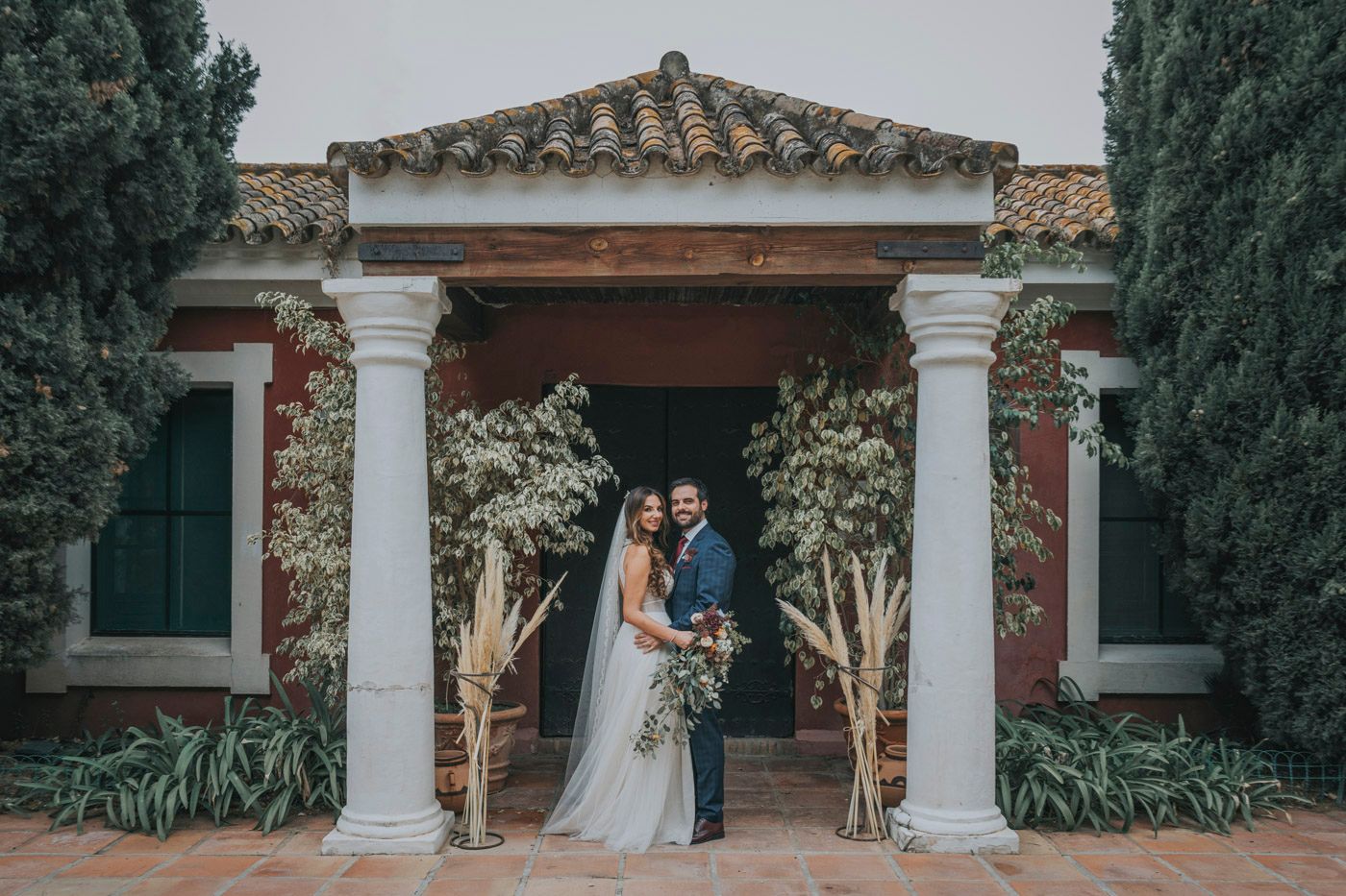 A wedding couple shares an intimate moment under a Mediterranean-style portico with white columns and greenery.