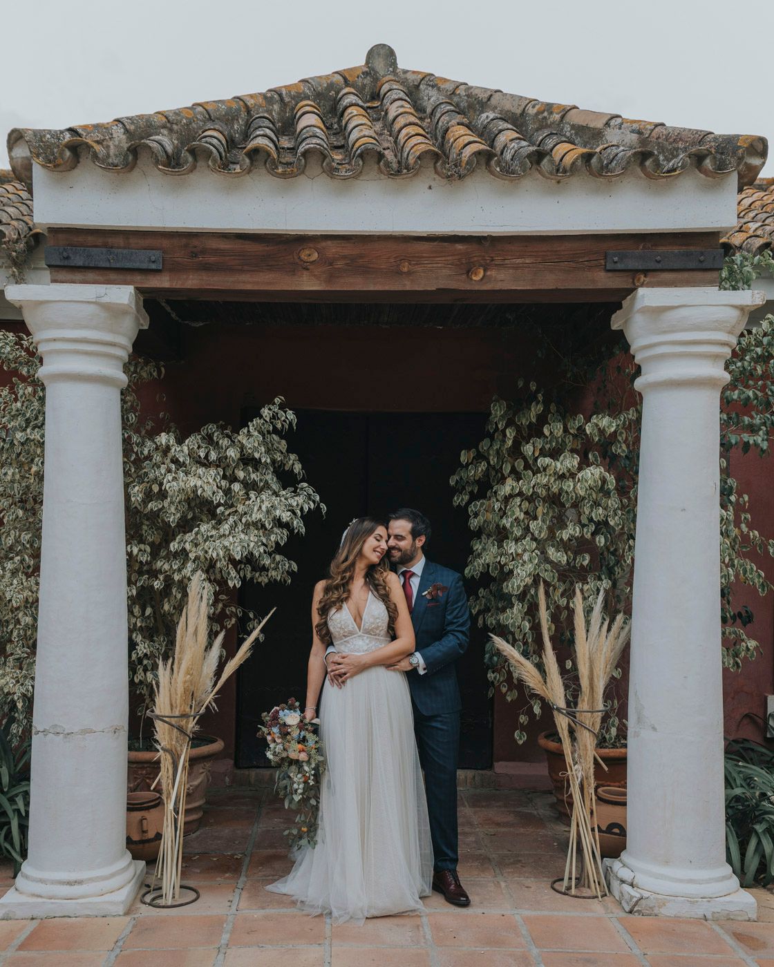 A romantic wedding portrait under an elegant Spanish-style portico with white columns and terracotta tiles.