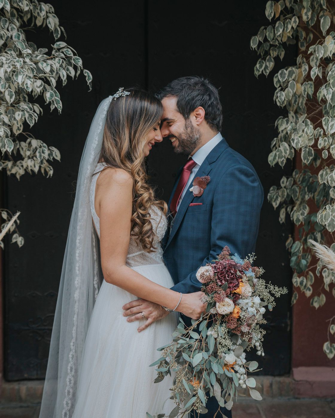 Romantic wedding couple embraces against dark wall with trailing vines and elegant floral bouquet.