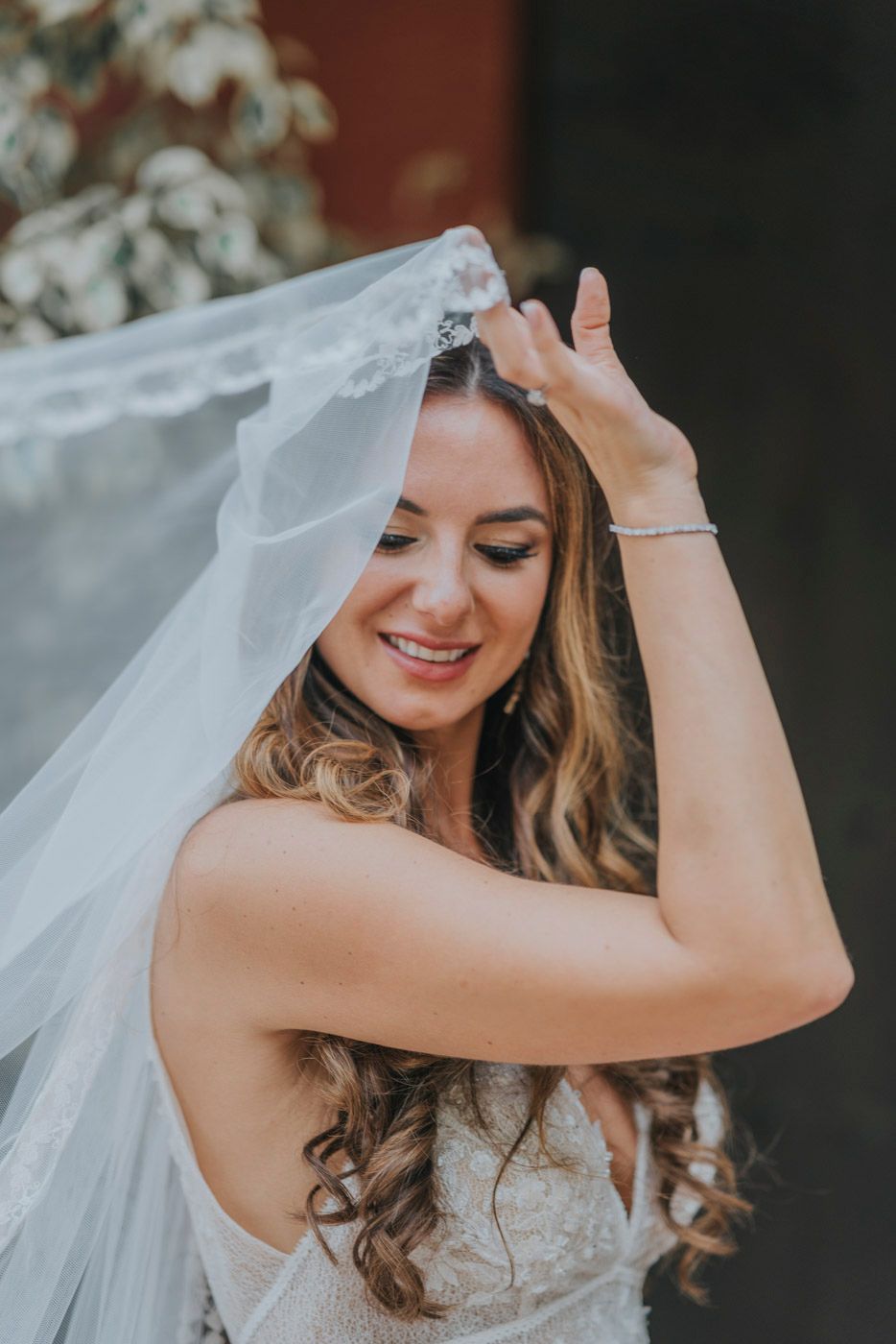 A bride in a white dress adjusts her flowing tulle veil against a dark backdrop.