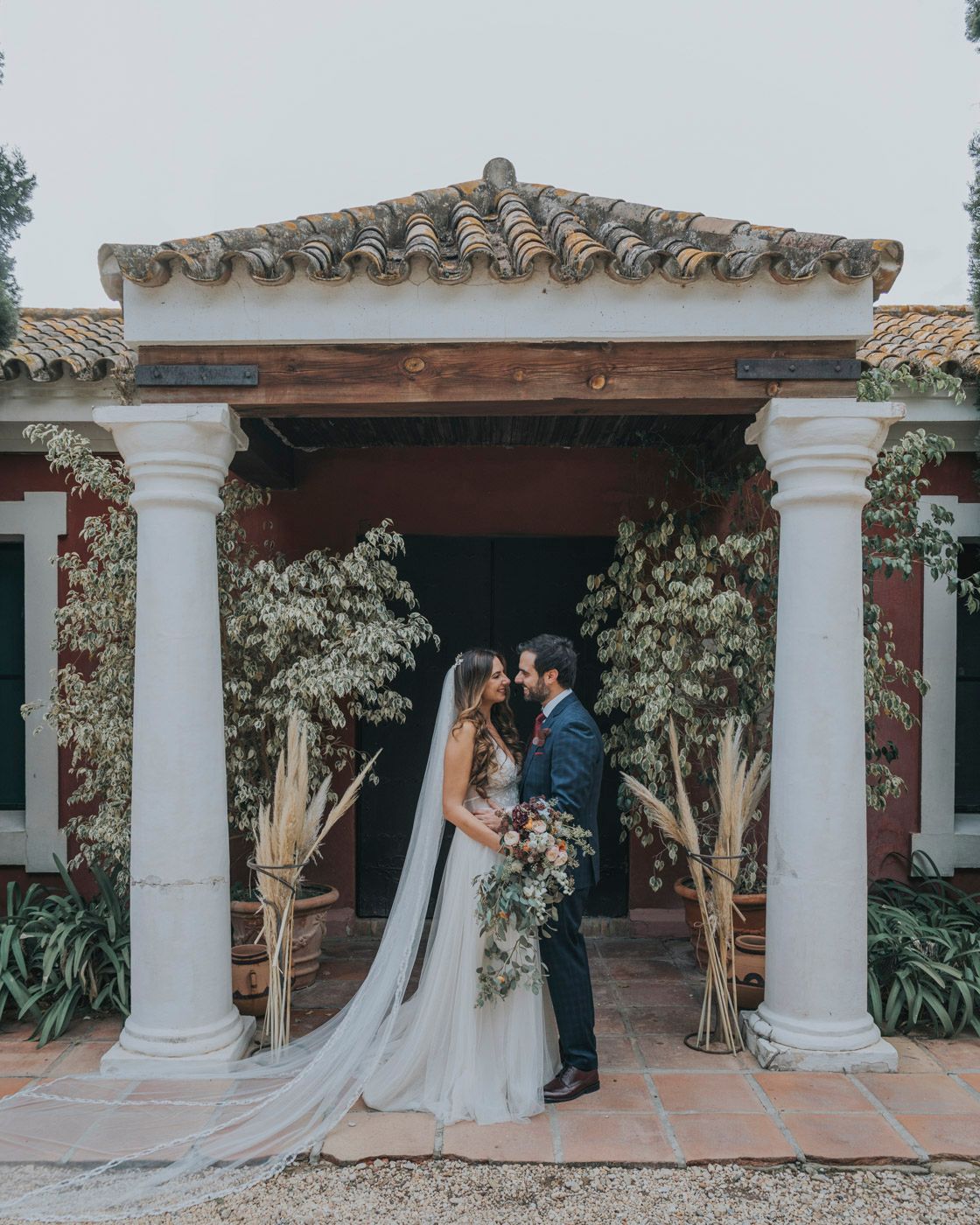 Newlyweds embrace under an ornate portico with white columns and trailing vines at a Mediterranean venue.