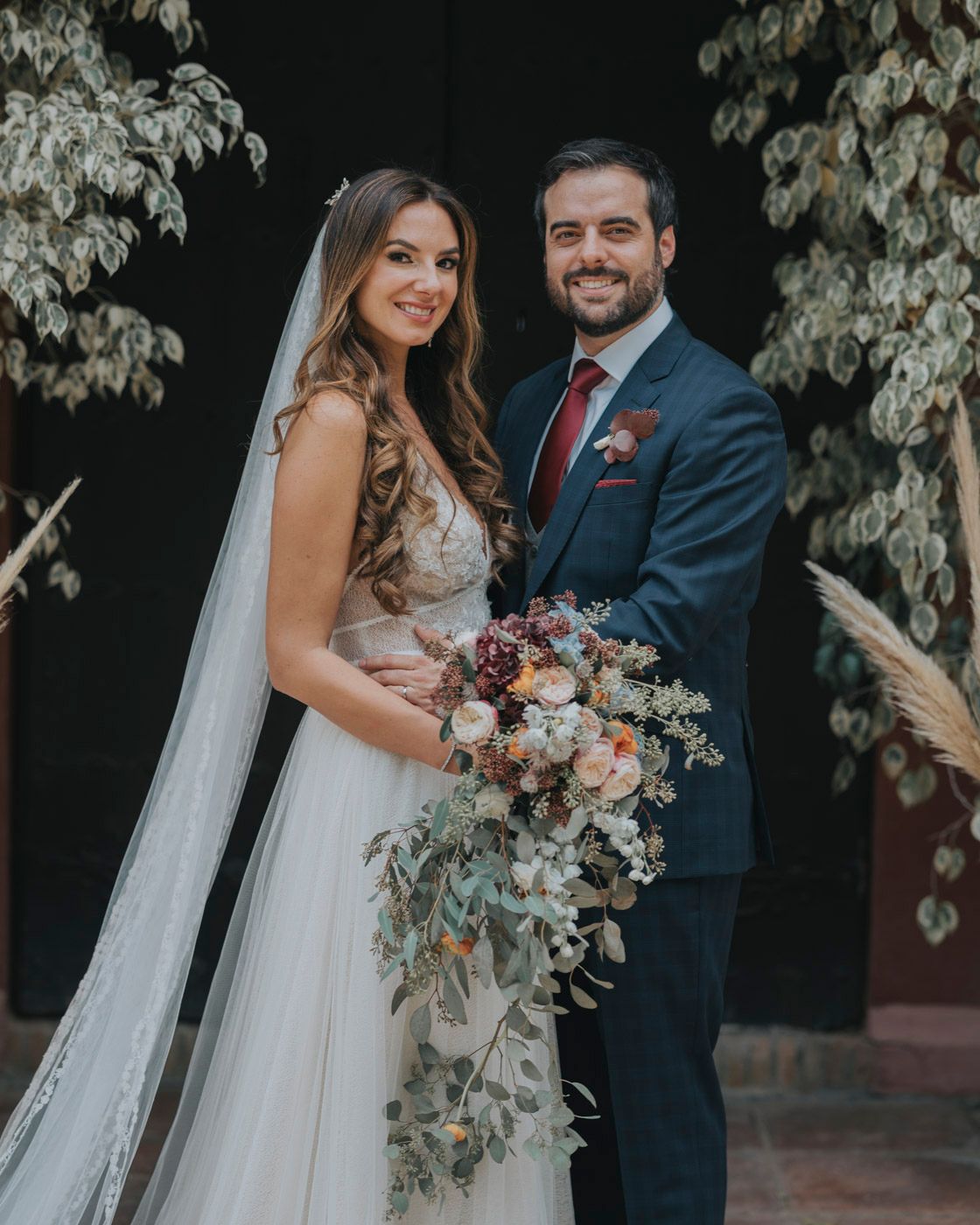 Wedding couple pose together outdoors with rustic floral decor and a cascading bouquet featuring burgundy and blush blooms.