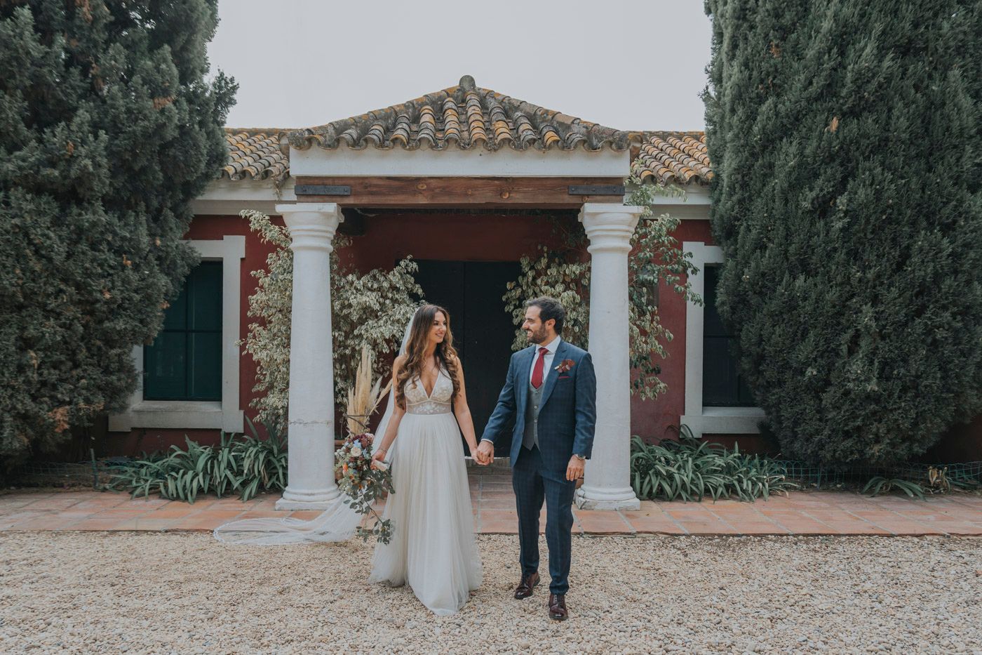 A couple poses on a Mediterranean-style villa entrance path framed by towering cypress trees and ornate columns.