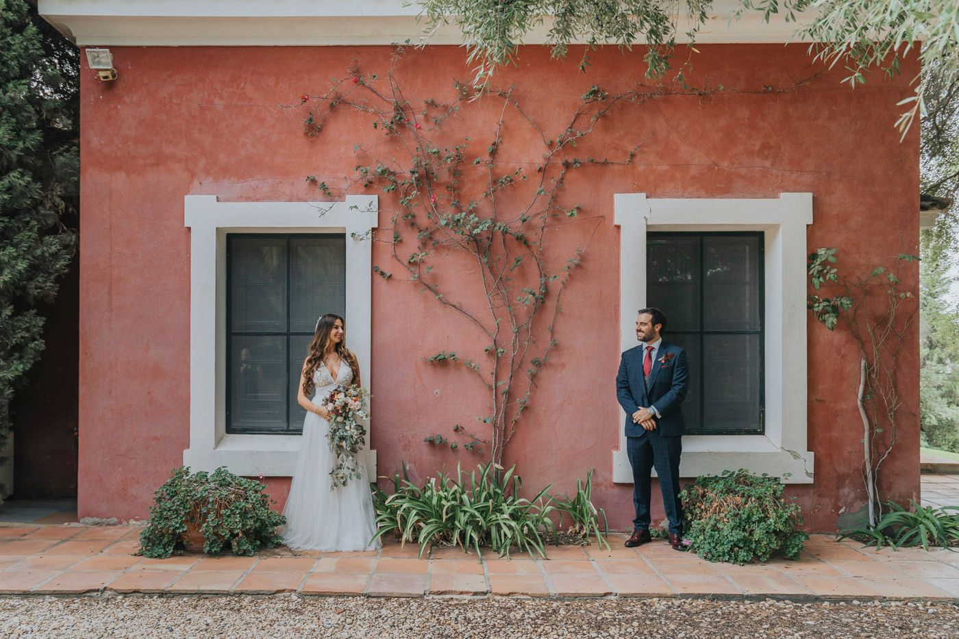 Couple in formal attire stands against coral-colored stucco wall with climbing vines and white-framed windows.