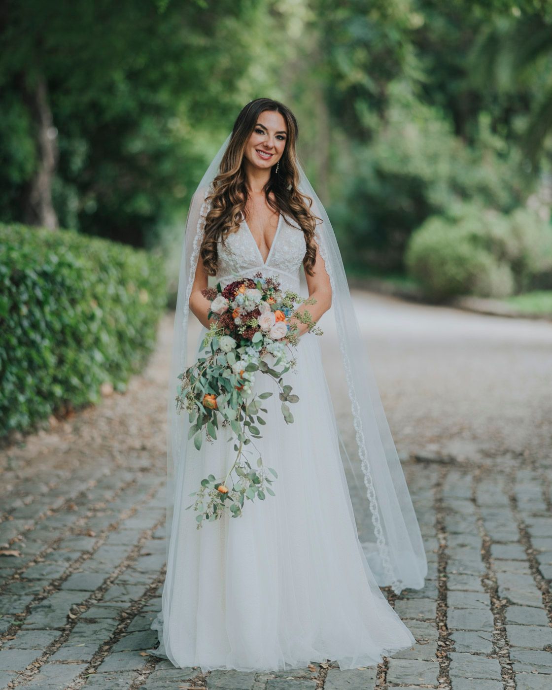 A bride in a flowing white tulle gown and long veil holds a cascading bouquet of flowers on a cobblestone path.